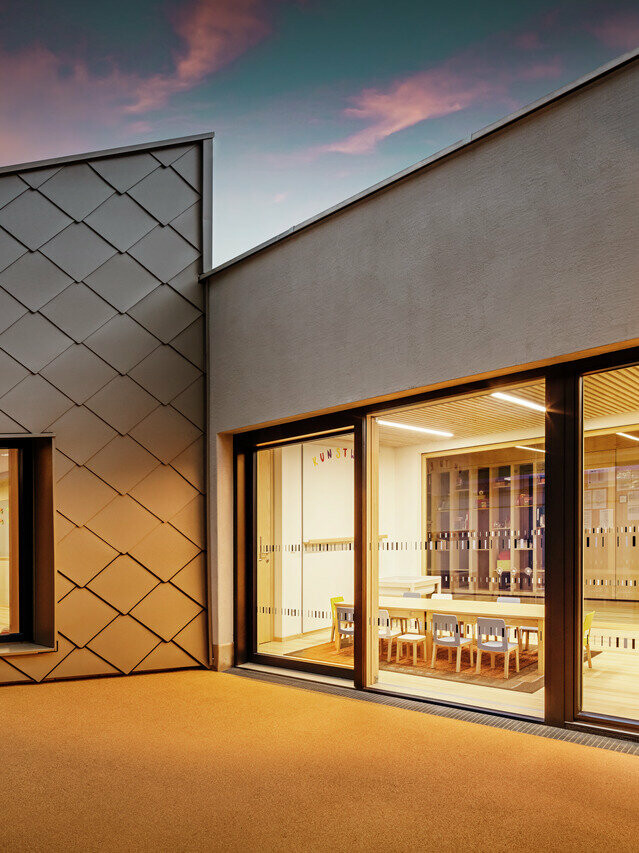 Abendansicht eines Teils des Schulungszentrums in Gerlos mit einer künstlerischen Fassade. Auf der linken Seite befindet sich die PREFA-Fenster Wandraute 44x44, während der Eingang auf der rechten Seite eine großzügige Fensterfront ist, die zu einem gut beleuchteten Innenraum mit Stühlen und Tischen führt. Die warme Innenbeleuchtung kontrastiert mit dem matten Licht der Abenddämmerung und unterstreicht die moderne Architektur und Gestaltung der Fassade.  Übersetzt mit DeepL.com (kostenlose Version)
