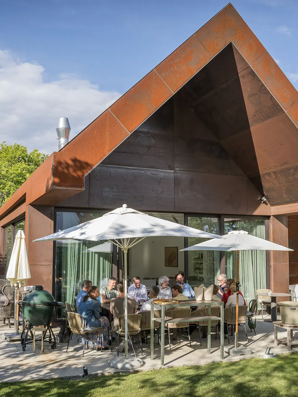 PREFARENZEN 2027 Expertendialog auf der Terrasse einer modernen Winzerresidenz: Jury-Mitglieder sitzen um einen großen Tisch, diskutieren intensiv über Architekturprojekte und blättern in Architekturbüchern. Die Szene wird von natürlichem Licht und grünem Garten umrahmt, während PREFA Aluminiumprodukte an Dach und Fassade des Gebäudes sichtbar sind. Das Bild vermittelt kreative Auseinandersetzung, konzentrierte Analyse und lebendigen Austausch. PREFA Dächer und Fassaden stehen für innovative, langlebige und ästhetisch anspruchsvolle Lösungen, die Design, Funktion und Inspiration in Architekturprojekten verbinden.