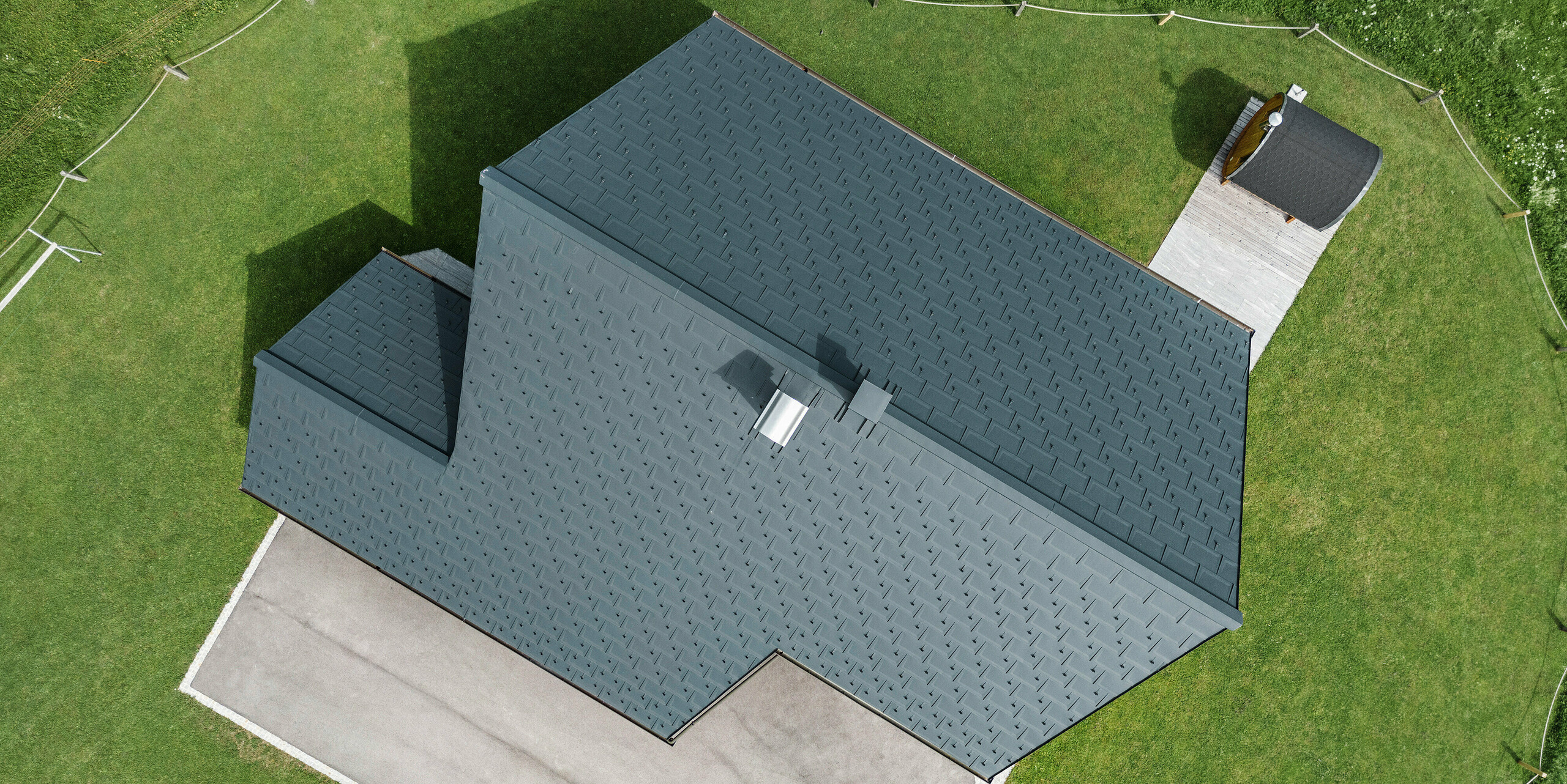 [Translate to Magyar:] Vogelperspektive auf ein Blockhaus in Gosau, Oberösterreich, mit einem präzise verlegten PREFA Aluminiumdach aus Dachplatten R.16 in P.10 Anthrazit. Das Bild demonstriert das robuste Dachsystem und geometrische Eleganz. Die Dachplatten R.16 verleihen dem traditionell österreichischen Holzhaus moderne Konturen, während die umliegenden grünen Grasflächen und die klar definierten Wege die saubere und gepflegte Landschaftsgestaltung hervorheben.