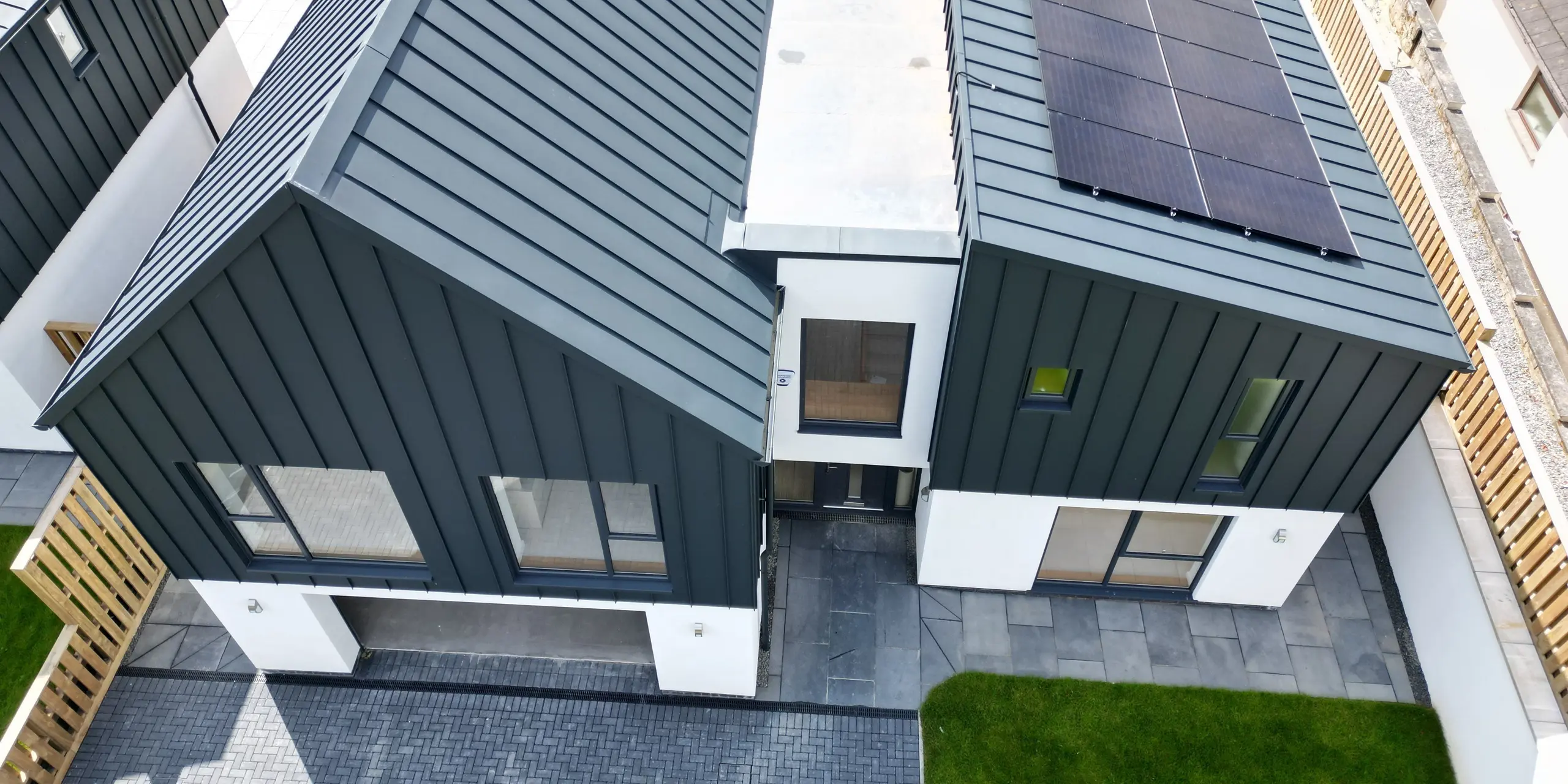Vogelperspektive auf ein Einfamilienhaus in Bridgerule, Devon, mit PREFALZ P.10 anthrazit als Stehfalzdeckung auf Dach und Fassade. Die moderne Architektur verbindet langlebiges Aluminium mit ästhetischer Langlebigkeit, ergänzt durch eine Solaranlage. Über der Garageneinfahrt thront ein Satteldach, der rechte Gebäudeteil erhielt ein Pultdach. Zwischen den Dächern verbindet eine Gebäudebrücke die beiden Baukörper.