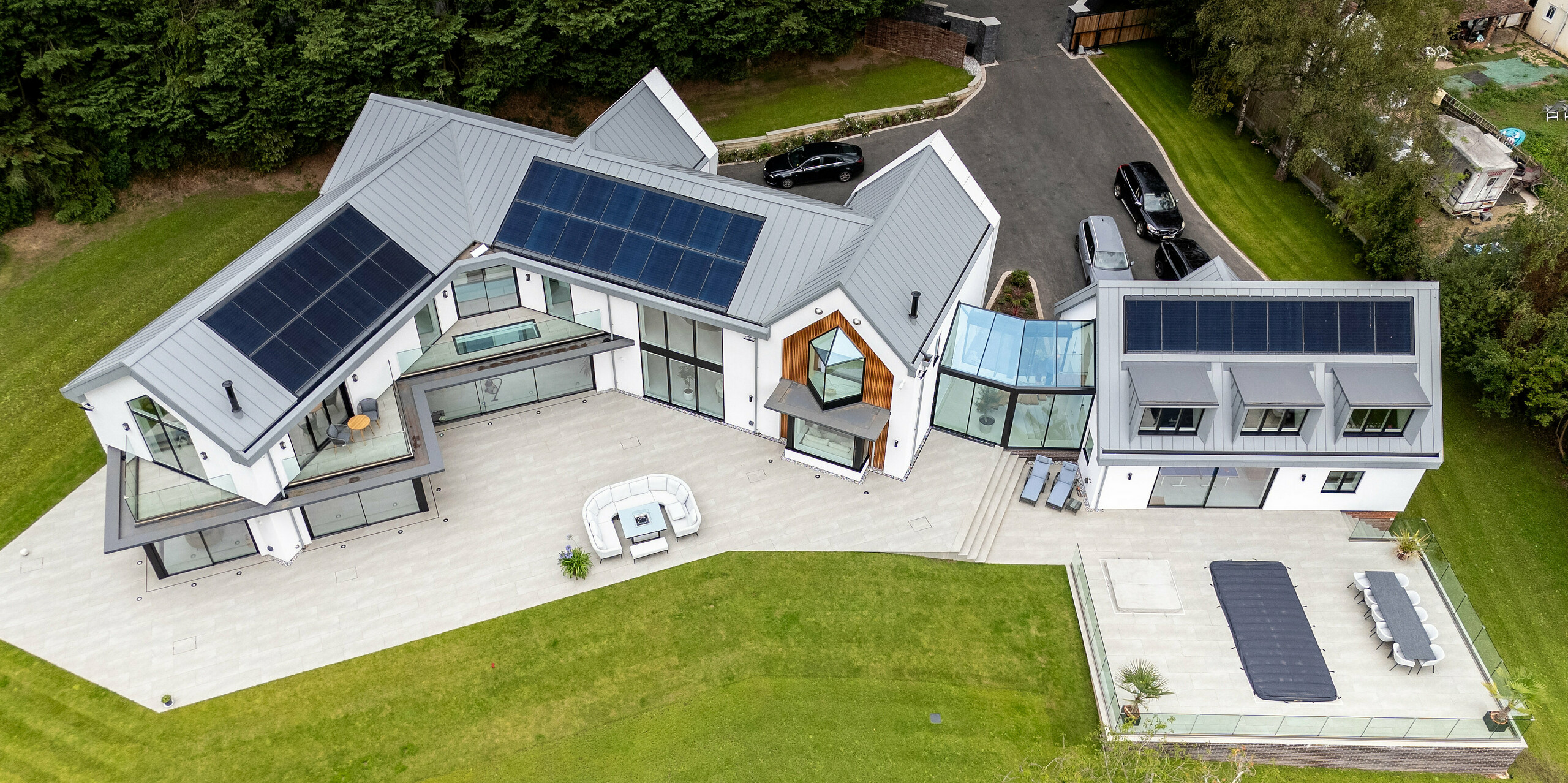 Das Bild zeigt eine beeindruckende Vogelperspektive der modernen Villa in Draycott in the Clay mit ihrem markanten PREFALZ-Dach in der Farbe Patinagrau. Die Stehfalzdeckung des Daches setzt sich harmonisch von der umliegenden grünen Landschaft ab und unterstreicht das moderne, elegante Design des Hauses. Die nahtlos in das Dach integrierten Solarpanels betonen die energieeffiziente Ausrichtung des Hauses. Die großzügige Terrasse und der gepflegte Garten vervollständigen das Bild dieser innovativen Architektur. Durch die Verwendung von PREFALZ ist höchste Qualität und Langlebigkeit garantiert.
