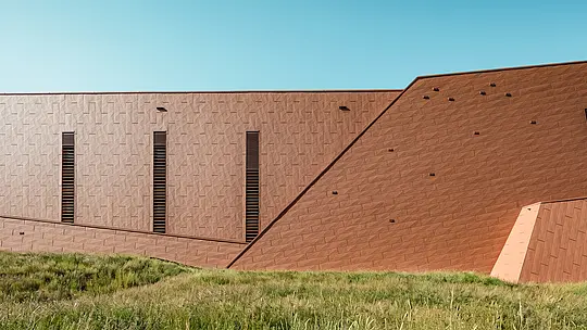 Das Trinkwasserpumpwerk in Lottum (Niederlande) fügt sich als skulpturales Bauvolumen harmonisch in die naturnahe Umgebung ein. Die prägnante PREFA Siding.X-Aluminiumfassade in warmer Rostoptik bildet einen spannenden Kontrast zu der grünen Wiese und den Hügeln und fasziniert mit ihrer fein strukturierten, lebendigen Oberfläche. Schlanke, vertikale Öffnungen setzen präzise Akzente. Die langlebigen, leichten und vollständig recycelbaren PREFA-Aluminium-Paneele überzeugen durch hohe Witterungsbeständigkeit, Farbkonstanz und eine nachhaltige, wartungsarme Gebäudehülle. Damit ist das Trinkwasserpumpwerk ein architektonischer Eyecatcher im Infrastrukturbau.