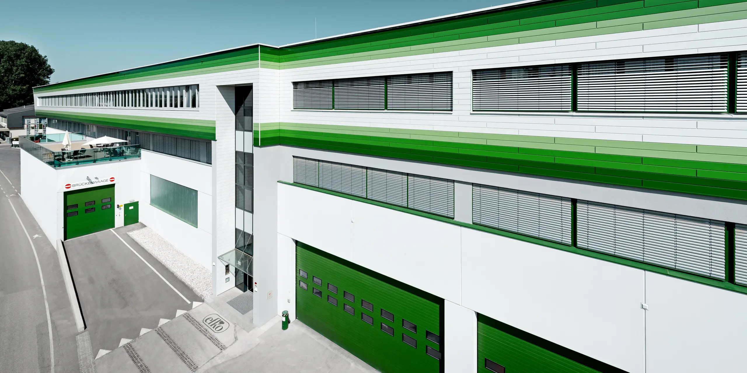Weitläufige Ansicht des Firmengebäudes der efko Frischfrucht und Delikatessen GmbH mit markanter PREFA Siding-Fassade in P.10 Prefaweiß und individuellen grünen RAL-Sonderbeschichtungen. Die horizontalen Aluminiumprofile betonen die außergewöhnliche Gebäudelänge und schaffen ein dynamisches Fassadenbild. PREFA Aluminium steht für langlebige Qualität, hohe Witterungsbeständigkeit, Farbstabilität und eine wartungsarme Lösung für moderne Unternehmensarchitektur.