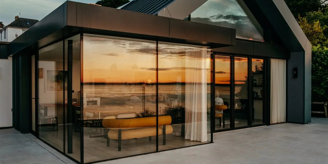 Nahaufnahme von der Terrasse eines luxuriösen Bungalows mit Meerblick auf der Isle of Wight. In den großzügigen Fenstern spiegelt sich der traumhafte Ausblick des Sonnenuntergangs an der Südküste Englands wider. Die großen Glasflächen sind umrahmt von PREFALZ in P.10 Dunkelgrau. Durch die Fenster sind Teile der modernen Innenarchitektur der Luxusimmobilie zu erkennen.