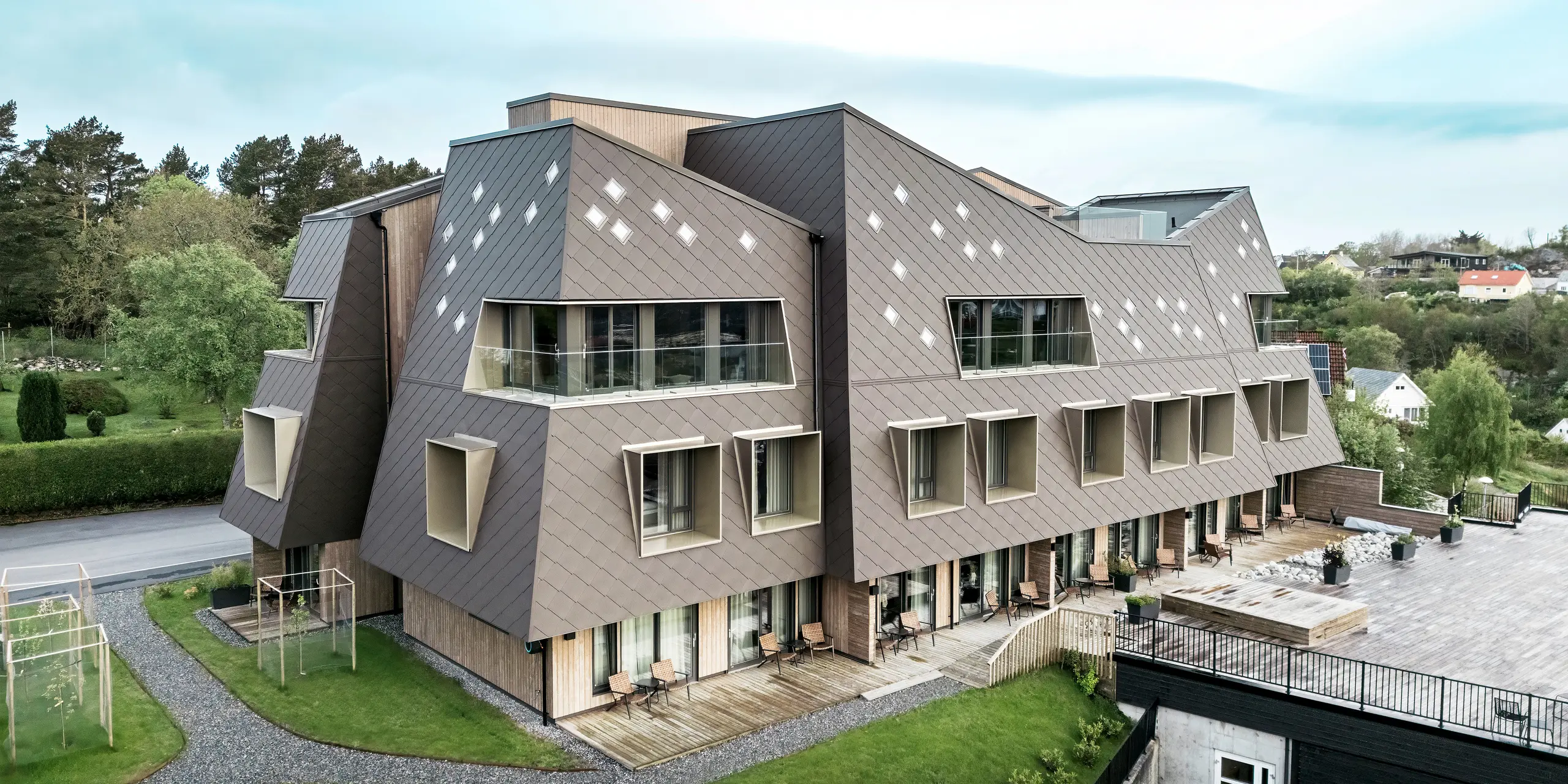 Außenansicht des Beckerwyc House in Bekkjarvik, Norwegen, mit Fokus auf die gepflegten Outdoor-Bereiche und die markante Fassade aus PREFA Wandrauten 44 × 44 in Nussbraun. Die skulpturale Architektur des Gebäudes, kombiniert mit einer großzügigen Terrasse und Balkonen, schafft eine harmonische Verbindung zwischen Innen- und Außenbereichen. Die futuristische Gestaltung und die natürliche Farbgebung passen sich gut in die umgebende Landschaft ein.