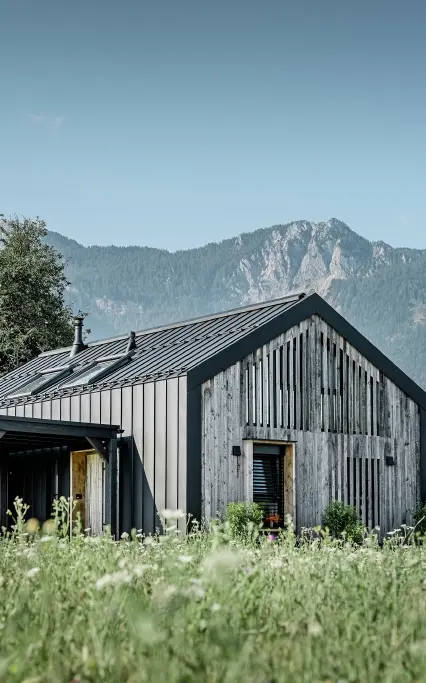 Moderner, funktionaler Wohnbau mit einer Kombination aus Holzfassade und PREFA Band- und Blechmaterial für das Dach. Die klare Architektur wird durch die elegante, metallische Dach- und Fassadenverkleidung unterstrichen, die ideal für anspruchsvolle Projekte ist. Das Bild veranschaulicht die vielseitigen Anwendungsmöglichkeiten der PREFA Produkte im Bereich Bänder und Bleche.