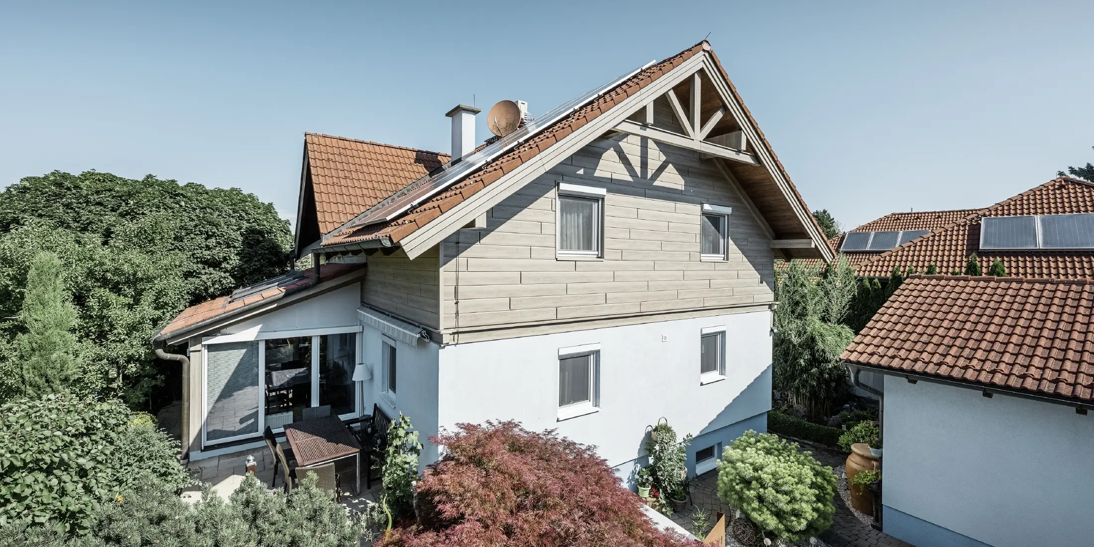 Családi ház tetővel és vörös tetőfedéssel, modern homlokzati kialakítással kombinálva. A felső oromfal PREFA bézs-szürke alumínium homlokzati panelekkel van burkolva, míg a földszint fehér vakolt homlokzattal rendelkezik. A tetőn napelemek találhatók. A ház egy zöld kertben fekszik, terasszal, kerti bútorokkal és buja növényzettel, hasonló tetőszerkezetű házakkal körülvéve.