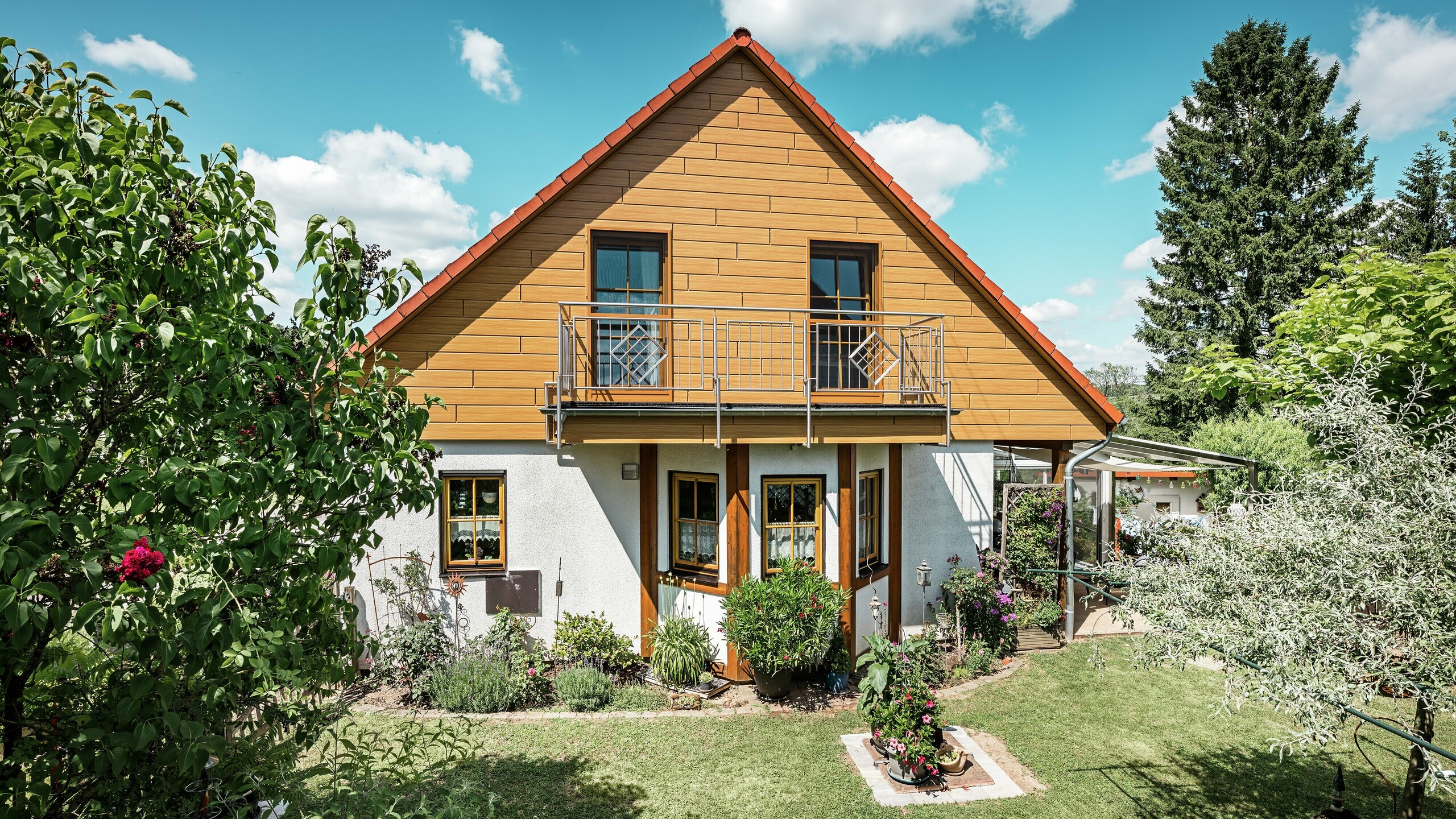 Ezen a képen a Blieskastelben található családi ház felújított oromfalának világos, természetes tölgy árnyalata ragyog a PREFA Sidingnek köszönhetően. A valósághű faoptikájú, kiváló minőségű alumínium homlokzat erőteljes vizuális akcentust ad, és lenyűgözően harmonizál a vörös cseréptetővel és a buja kerttel. Virágok, cserjék és dús növényzet veszi körül, így a ház barátságos és gondozott benyomást kelt. A PREFA felület ötvözi a természetes megjelenést olyan funkcionális előnyökkel, mint a magas időjárásállóság, a stabilitás és a szinte karbantartásmentes tartósság – egy esztétikai csúcspont, amely fenntartható hozzáadott értéket képvisel.