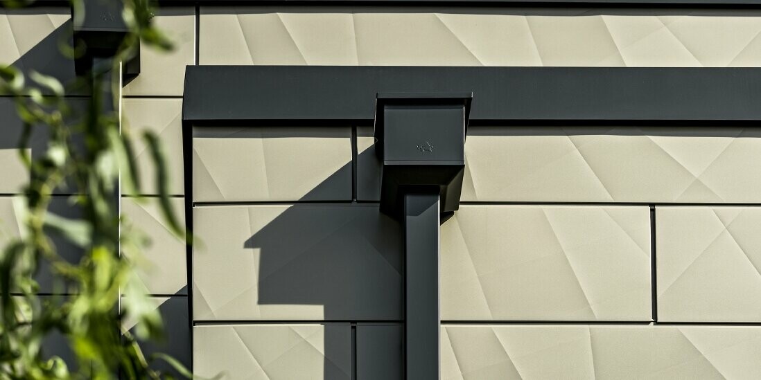 Modern, lapostetős családi ház keskeny ablakokkal. A homlokzat burkolata az egyedi megjelenésű, bronzszínű PREFA Siding.X homlokzati elemekből készült. A csatlakozások antracit színűek. Tető-vízelvezetés céljára a PREFA szögletes lefolyócső szolgál, a hozzá megfelelően illeszkedő antracit színű vízgyűjtő üsttel.