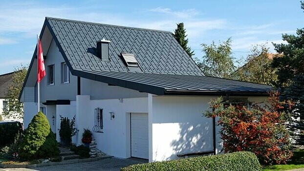Nyeregtetős felújított ház és hozzá tartozó garázs. A tetőt PREFA Classic elem, a garázst antracit színű Prefalz borítja. A ház előtt zászlórúd svájci zászlóval.