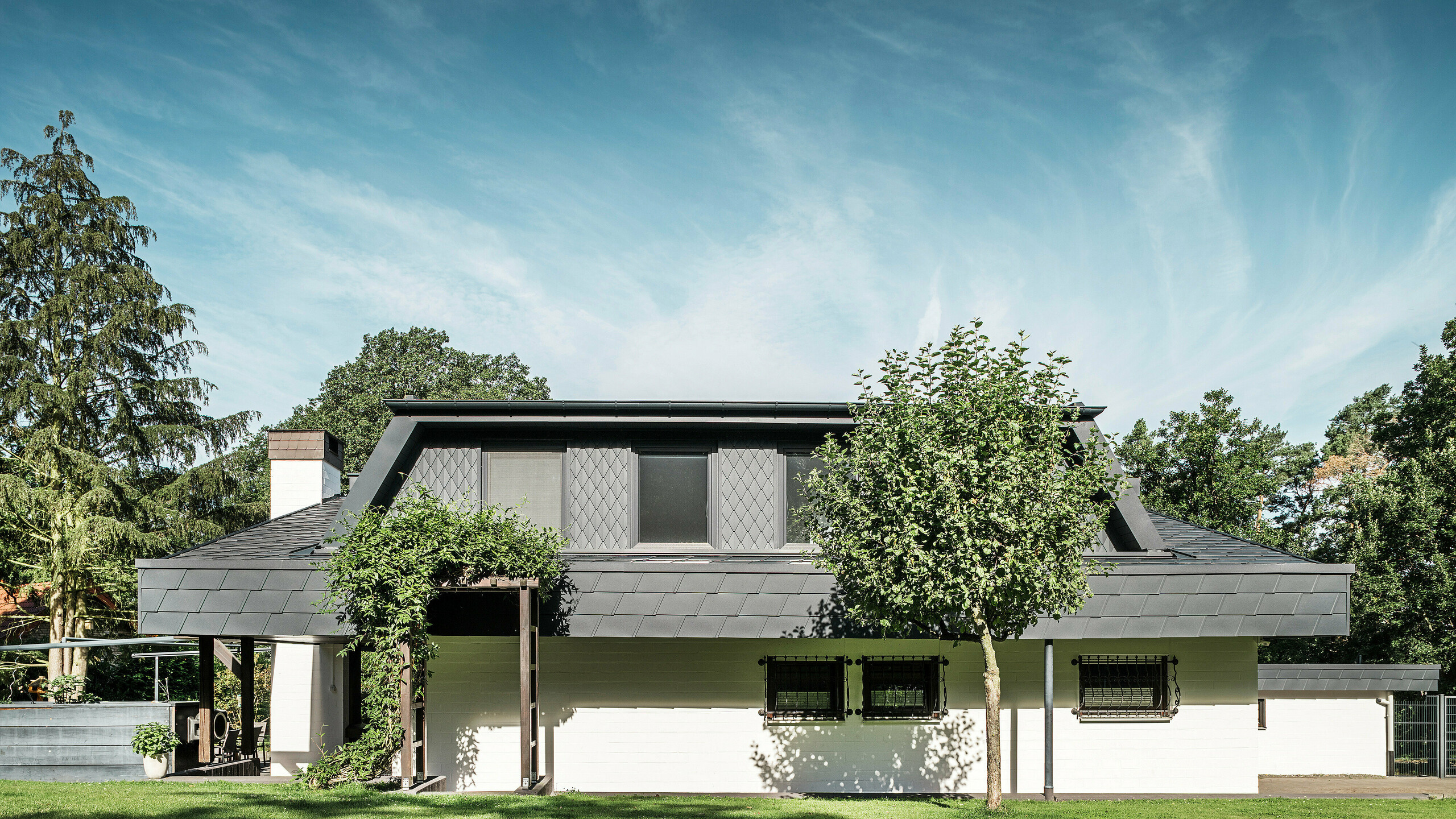 Egy mechernichi családi ház kert felőli homlokzata, nagy trapéz alakú tetőablakkal, amelyet apró, antracit színű PREFA P.10 tetőfedő rombuszok borítanak. A tetőt ugyanebben a színárnyalatban PREFA DS.19 tetőfedő zsindelyek borítják. Az alsó szint homlokzata fehérre van festve, és egy növényzettel borított pergola teszi teljessé a kertben kialakult összképet. A fák és cserjék keretezik a kilátást, és gondoskodnak a természetbe való harmonikus beilleszkedésről.