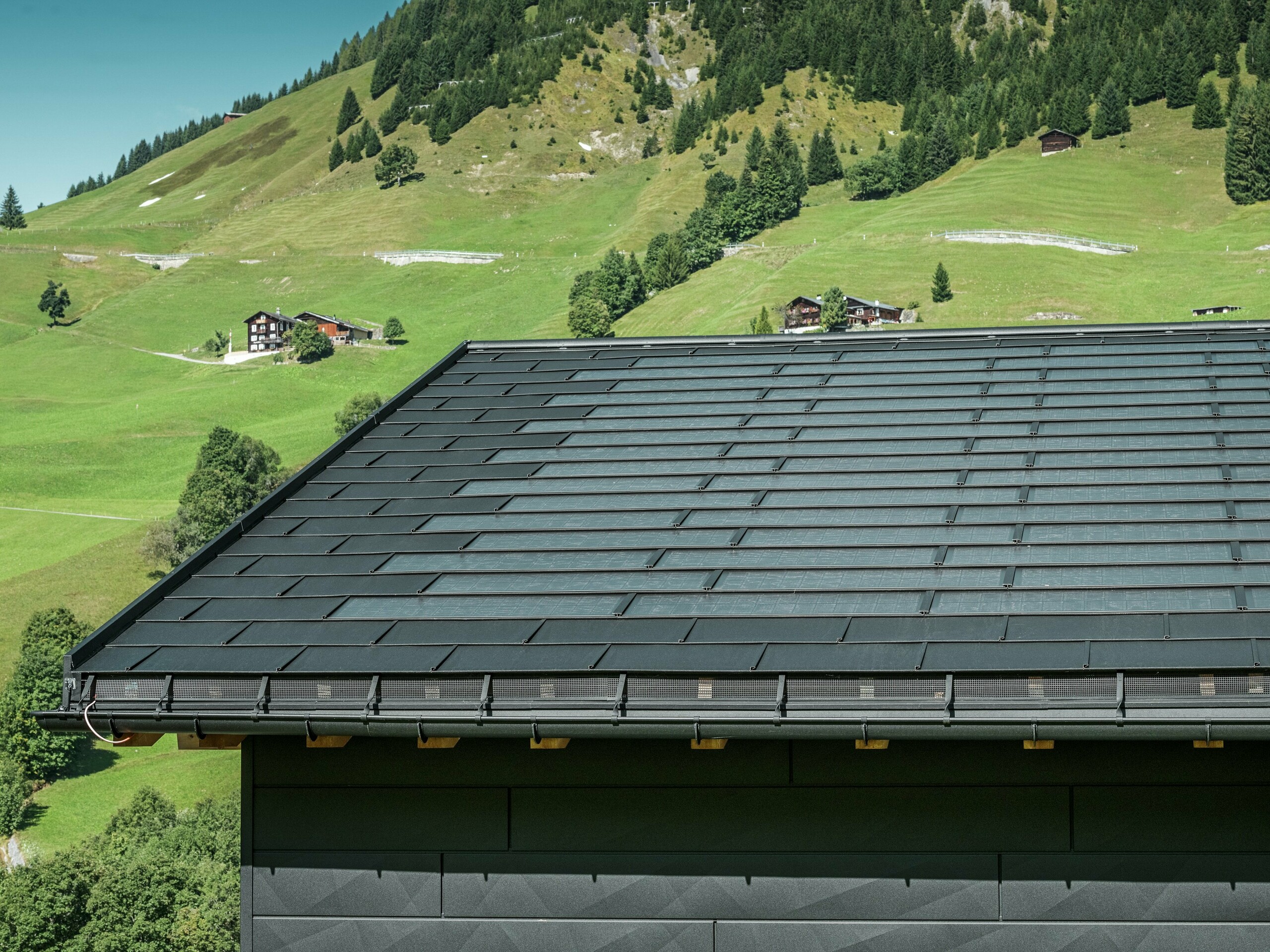 Közelkép egy modern családi ház ereszéről Svájc alpesi környezetében, Ascharinában. A tetőt az innovatív PREFA Solardachplatte és a hagyományos R.16-os Classic elem borítja, mindkettő P.10 fekete színben. A PREFA Hängerinne és a PREFA Ablaufrohr biztosítja a tető megbízható vízelvezetését. Az alsó képen a jellegzetes PREFA Siding.X homlokzati panelek láthatók. Az alumínium termékek tiszta vonalvezetése hangsúlyozza a modern dizájnt a festői hegyvidéki táj közepette.