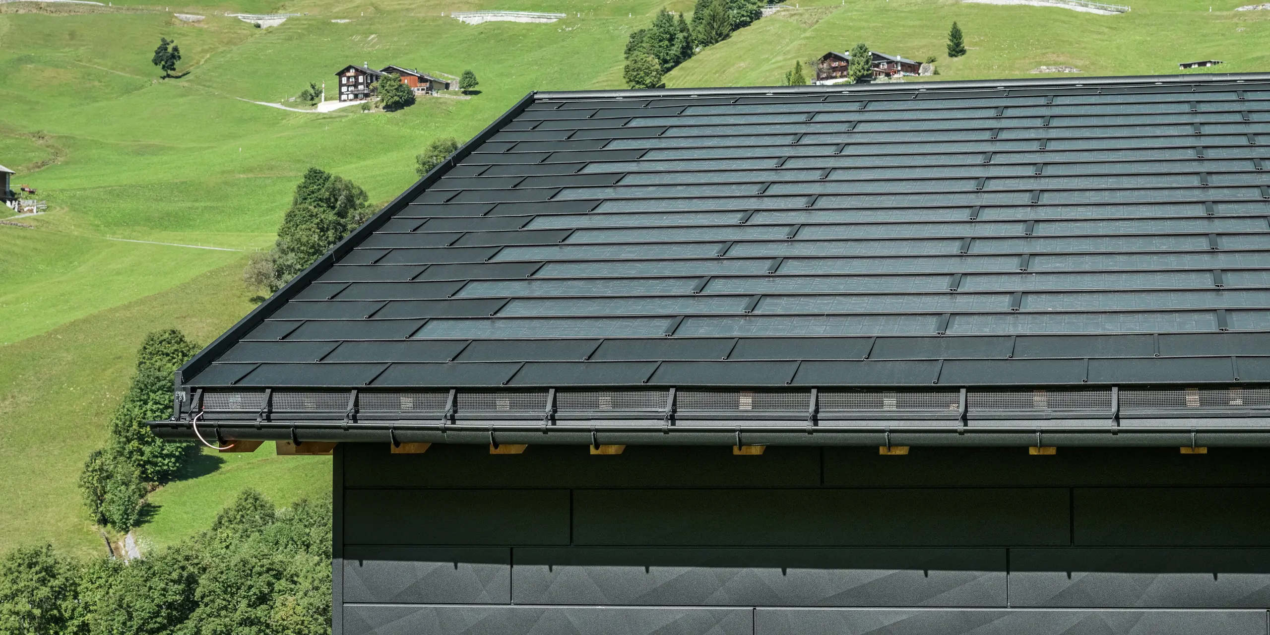 Közelkép egy modern családi ház ereszéről Svájc alpesi környezetében, Ascharinában. A tetőt az innovatív PREFA Solardachplatte és a hagyományos R.16-os Classic elem borítja, mindkettő P.10 fekete színben. A PREFA Hängerinne és a PREFA Ablaufrohr biztosítja a tető megbízható vízelvezetését. Az alsó képen a jellegzetes PREFA Siding.X homlokzati panelek láthatók. Az alumínium termékek tiszta vonalvezetése hangsúlyozza a modern dizájnt a festői hegyvidéki táj közepette.