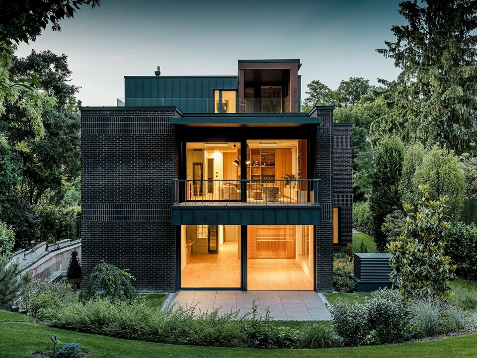Die Aufnahme zeigt die moderne Rückseite der Budapester Mehrgenerationen‑Villa in der Abenddämmerung. Großzügige Glasfronten öffnen den Blick auf warm erleuchtete Innenräume, während die Kombination aus dunkelroter Backsteinfassade und PREFALZ Aluminium in P.10 Anthrazit klare architektonische Linien betont. Die präzise ausgeführten Winkelstehfalze unterstreichen die hochwertige Verarbeitung und Langlebigkeit der PREFA Aluminiumprodukte. Umgeben von üppigem Grün fügt sich das Gebäude harmonisch in die Gartenlandschaft ein und vermittelt einen eleganten, modernen Charakter.