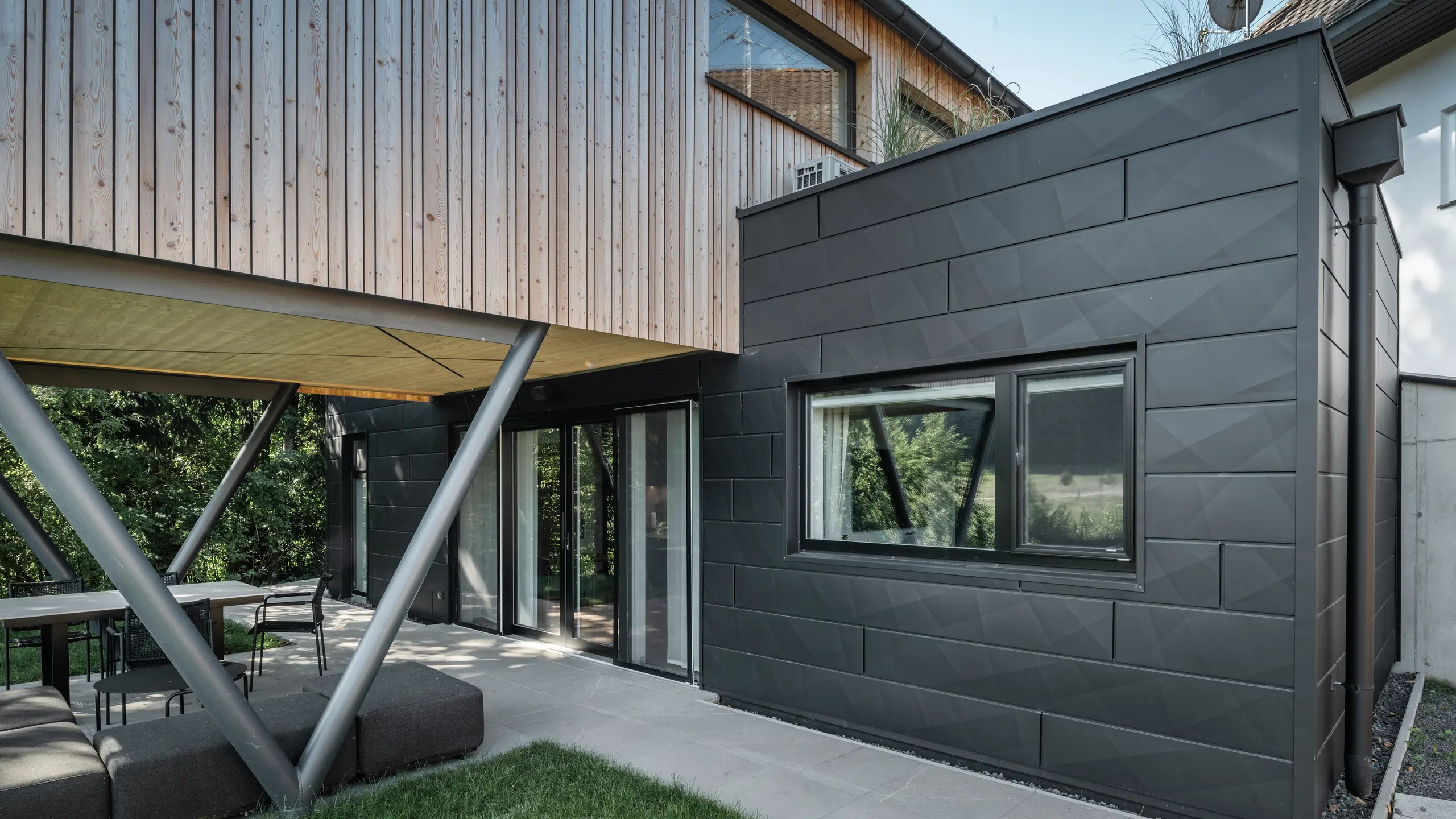Építészeti kiemelkedő: „Woodstock Chalet 5” St. Martin am Innkreisban, Felső-Ausztriában. A modern esküvői szálláshelyet antracit színű PREFA Siding.X homlokzati panelek használata emeli ki. Az alumínium elemek erős kontrasztot képeznek a világos fa panelekkel és a fém tartószerkezetekkel. A sötét ablakkeretek harmonikusan illeszkednek a homlokzat kialakításába, ami elegáns megjelenést kölcsönöz az épületnek. Ez a kép a természetes fa textúrája és az alumínium homlokzat ipari varázsa közötti esztétikai egyensúlyt mutatja, amely a kortárs épületek kialakítását jellemzi.