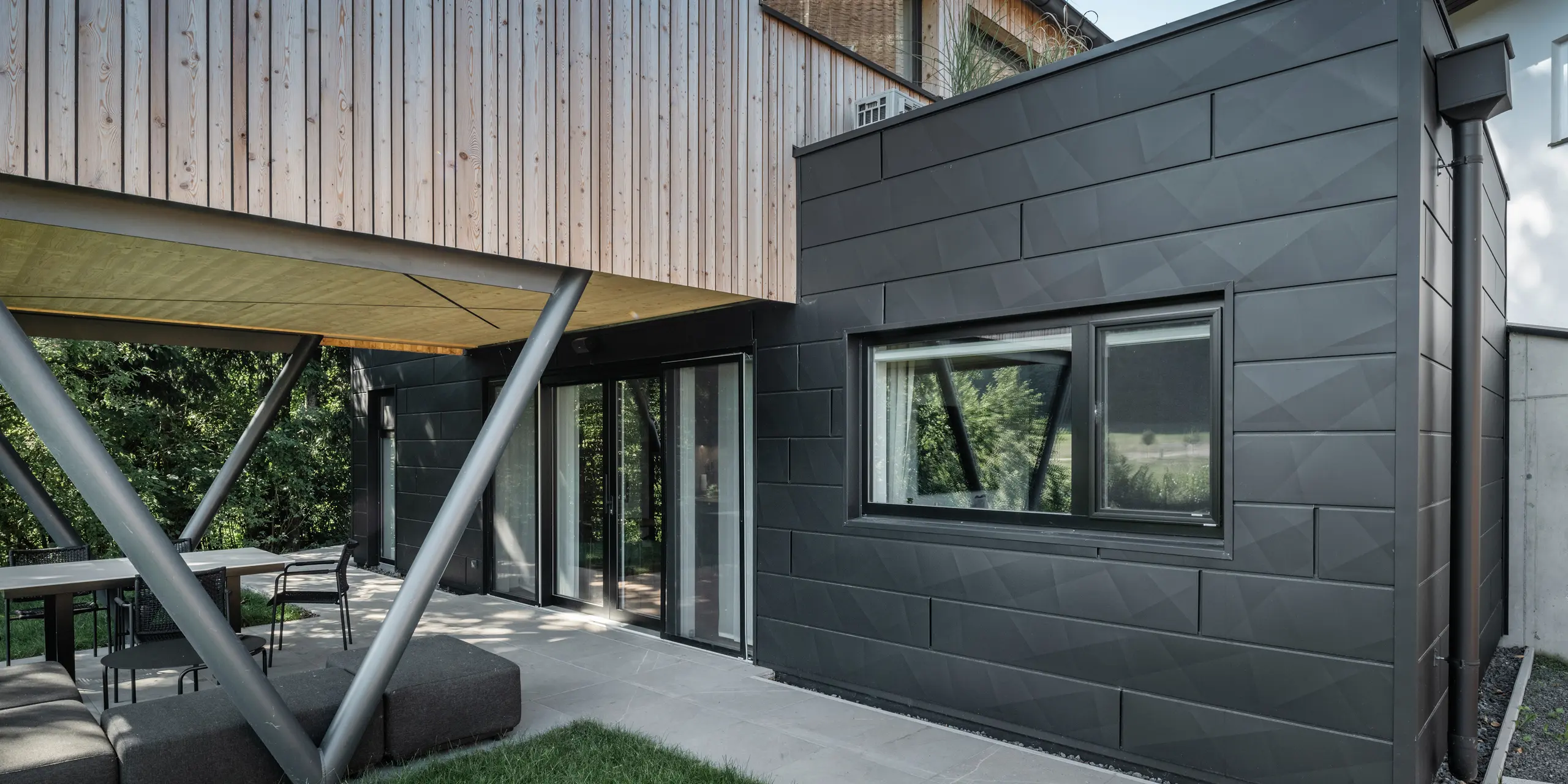 Építészeti kiemelkedő: „Woodstock Chalet 5” St. Martin am Innkreisban, Felső-Ausztriában. A modern esküvői szálláshelyet antracit színű PREFA Siding.X homlokzati panelek használata emeli ki. Az alumínium elemek erős kontrasztot képeznek a világos fa panelekkel és a fém tartószerkezetekkel. A sötét ablakkeretek harmonikusan illeszkednek a homlokzat kialakításába, ami elegáns megjelenést kölcsönöz az épületnek. Ez a kép a természetes fa textúrája és az alumínium homlokzat ipari varázsa közötti esztétikai egyensúlyt mutatja, amely a kortárs épületek kialakítását jellemzi.