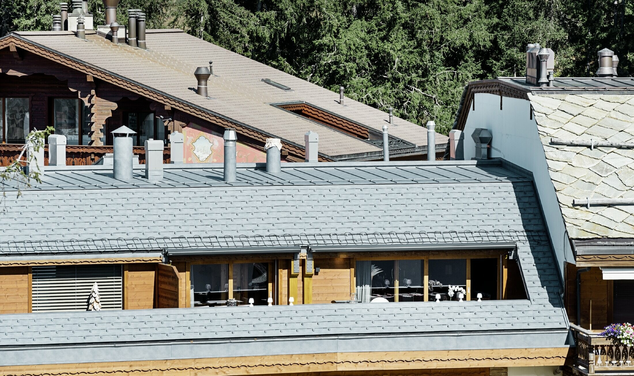 Társasház Crans-Montana településen, hegyekkel a háttérben, játékos fa homlokzattal és márványszürke PREFA alumínium tetőfedő zsindely fedéssel