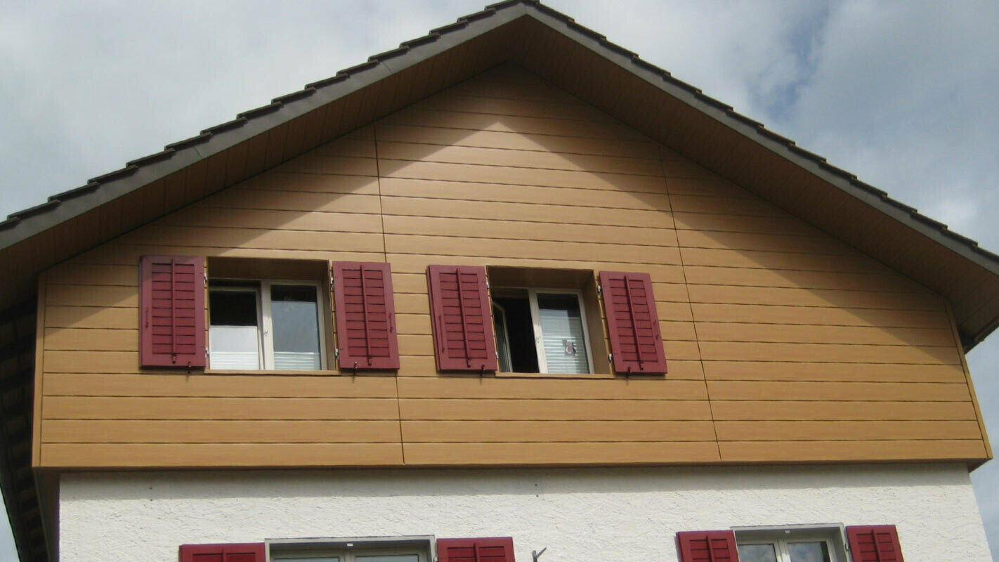 Ház fa hatású homlokzata, vízszintesen elhelyezett PREFA Siding elemek, piros zsalugáterek