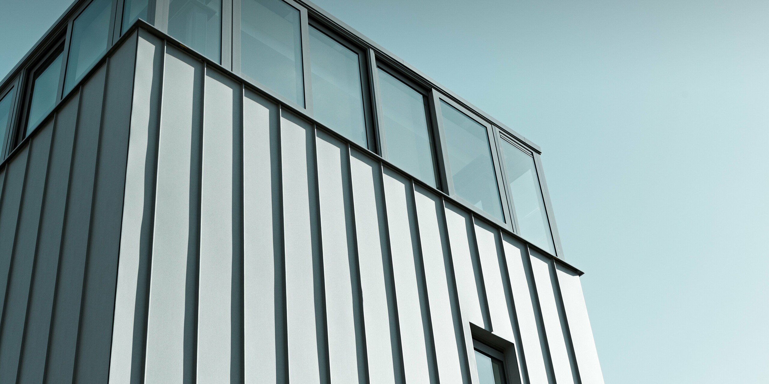 Dynamische Detailaufnahme einer modernen PREFALZ Fassade in Patinagrau. Die klare Linienführung der vertikalen Stehfalzbahnen trifft auf großzügige Verglasung im Obergeschoss, die Licht und Transparenz schafft. Die langlebige PREFA Aluminiumfassade sorgt für dauerhaften Schutz und setzt architektonisch ein markantes Statement.