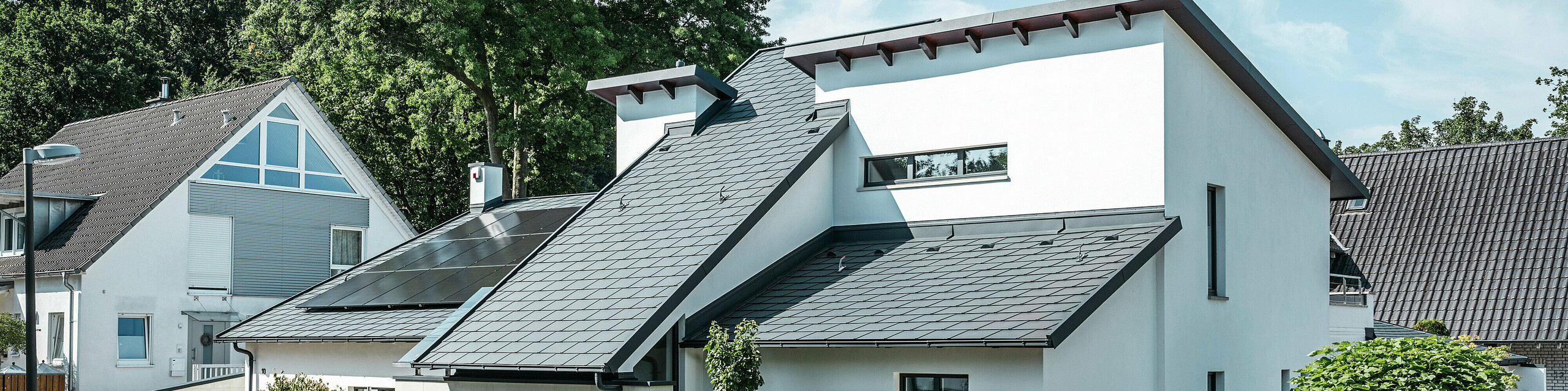 Modern családi ház fehér vakolt homlokzattal és sötét PREFA alumínium tetővel. A tetőn integrált fotovoltaikus modulok és antracit színű alumínium tető-vízelvezetés rendszer látható. Az épület egy gondozott lakótelepen található, virágos előkertekkel, burkolt bekötőúttal és környező családi házakkal.