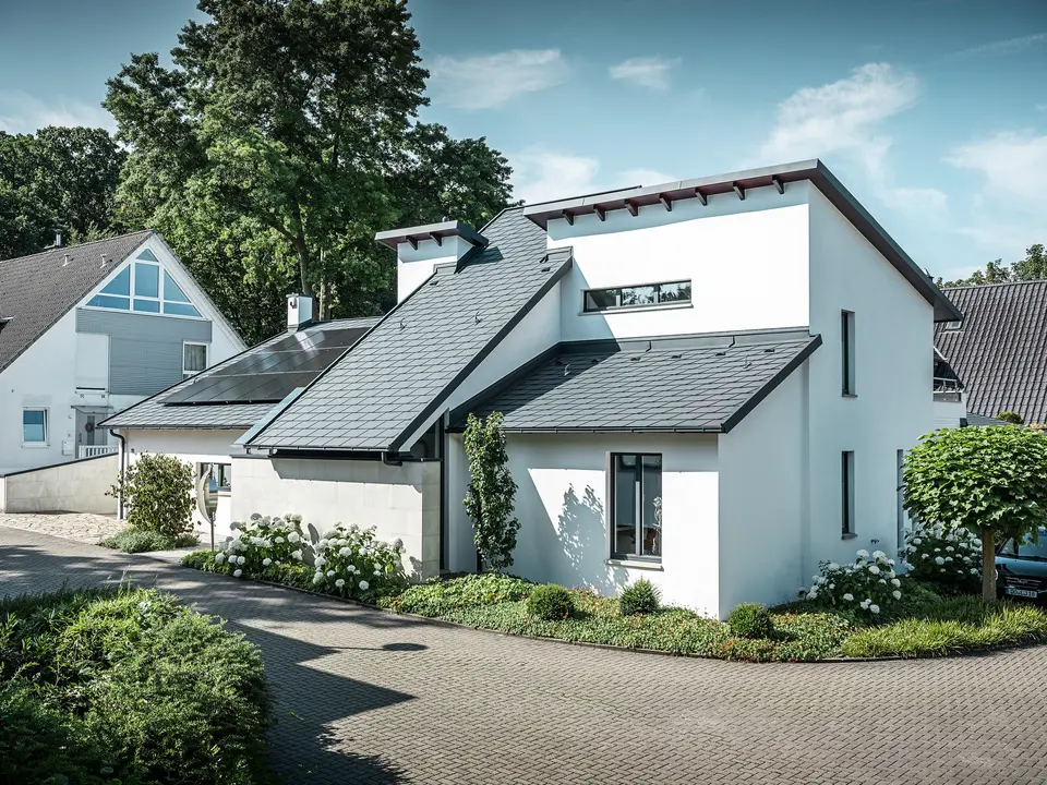Modern családi ház fehér vakolt homlokzattal és sötét PREFA alumínium tetővel. A tetőn integrált fotovoltaikus modulok és antracit színű alumínium tető-vízelvezetés rendszer látható. Az épület egy gondozott lakótelepen található, virágos előkertekkel, burkolt bekötőúttal és környező családi házakkal.
