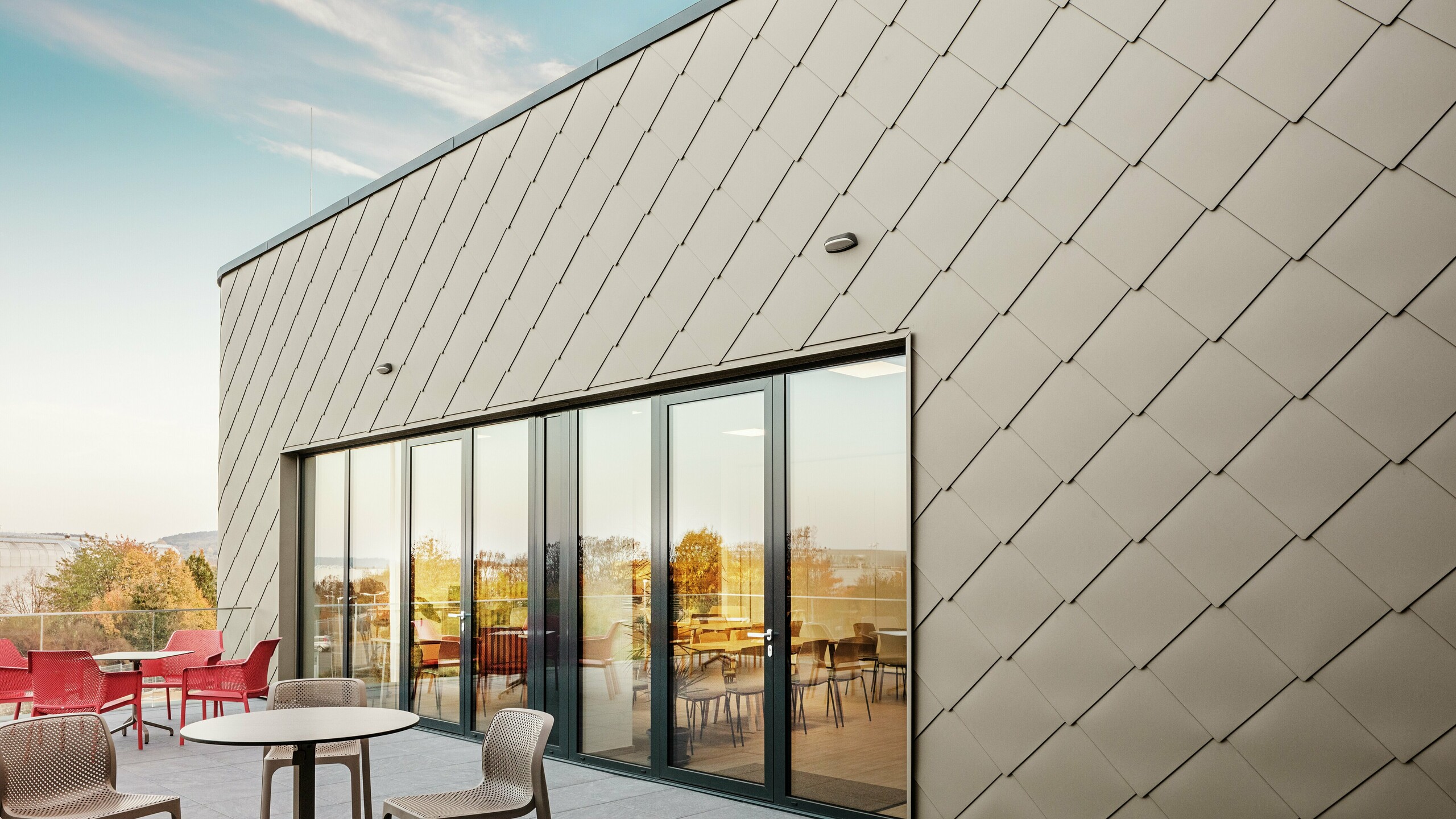 Die Fassade des neuen PREFA Standortes in Törökbálint besticht durch die eleganten PREFA Wandrauten 44 × 44 in Bronze. Die präzise verlegte Aluminiumfassade sorgt für ein modernes, dynamisches Erscheinungsbild und bietet gleichzeitig maximalen Schutz vor Witterungseinflüssen. Die großzügige Glasfront verbindet Innen- und Außenbereich und schafft auf der großzügigen Terrasse eine helle und einladende Atmosphäre.