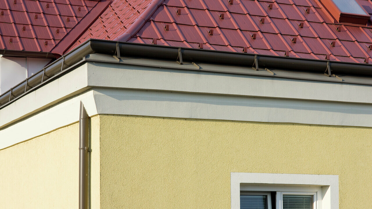 Házsarok sárgára vakolt homlokzattal és barna PREFA fekvő ereszcsatornával (zámcsatorna) piros PREFA Classic elem tetővel