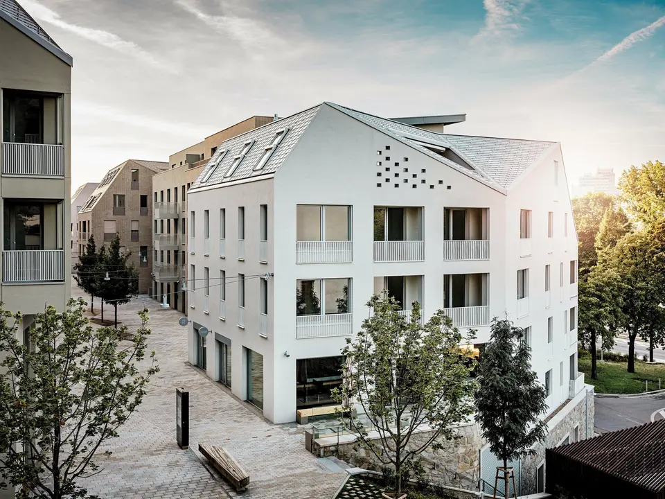 Modernes Wohnquartier Vydrica in Bratislava mit klarer Architektursprache und ruhiger Gassenatmosphäre: Die steil geneigten Satteldächer mit markanten Dachfenstern sind mit PREFA Aluminium Dachschindeln in P.10 Prefaweiß, Bronze und P.10 Sandbraun gedeckt und bilden einen eleganten Eyecatcher im Sonnenlicht. Die präzise ausgeführten Linien und homogenen Flächen unterstreichen die hochwertige Gestaltung. PREFA Aluminium überzeugt durch Langlebigkeit, geringes Gewicht und hohe Witterungsbeständigkeit – ideal für komplexe Dachformen und eine nachhaltige, wartungsarme Gebäudehülle.