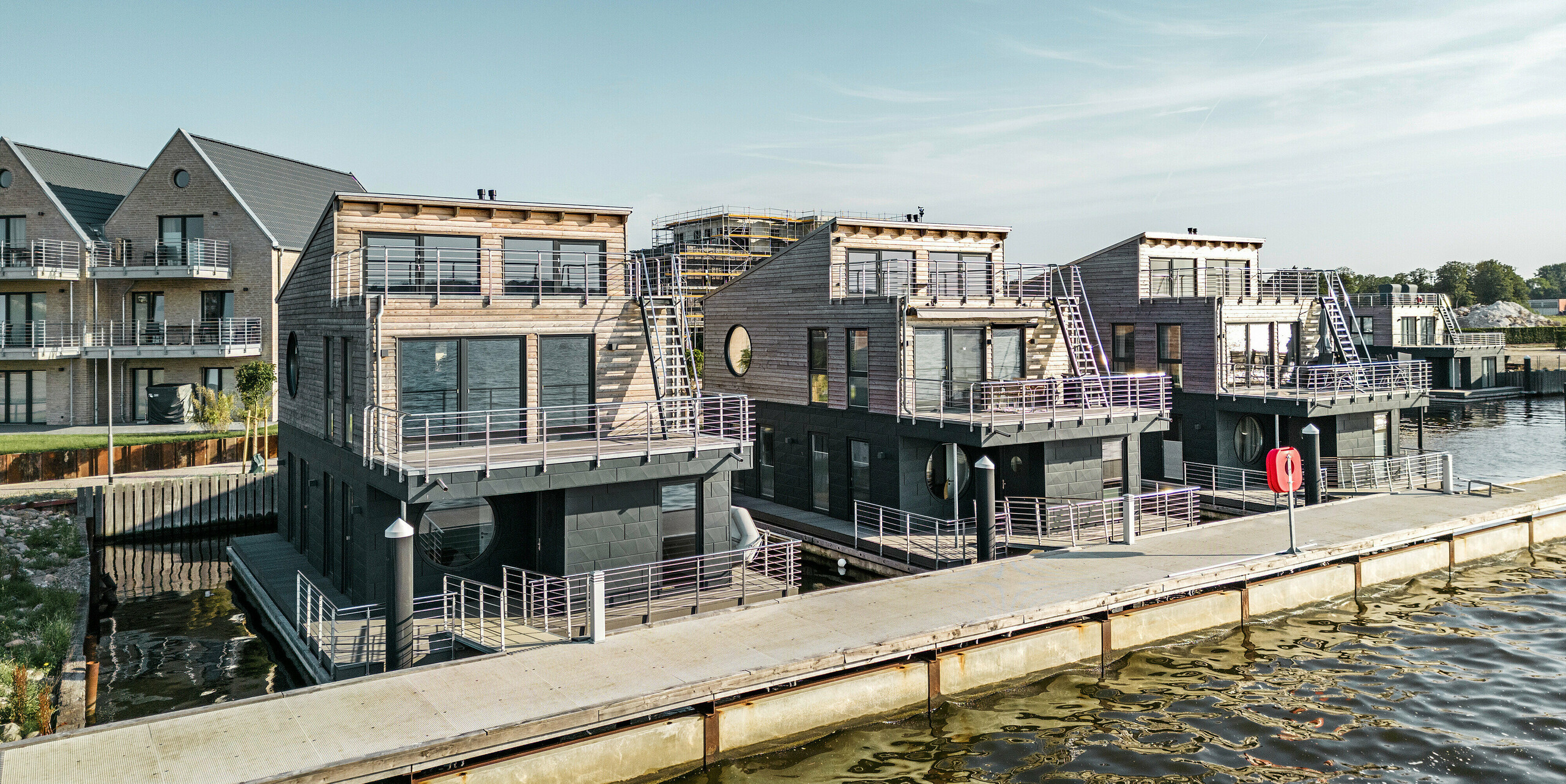 Einzigartige Wasserhaus-Komposition im Pionierhafen Schleswig, wo modernes Wohnen auf dem Wasser mit PREFA Siding.X in P.10 Anthrazit realisiert wird. Die Fassaden der Häuser kombinieren natürliche Holzelemente mit robusten Aluminiumpaneelen, die sich nahtlos in die Küstenlandschaft einfügen. Die Architektur reflektiert einen maritimen Lifestyle, begünstigt durch die direkte Lage am Wasser und unterstrichen durch die hochwertige, korrosionsbeständige Verkleidung, die Schutz und Ästhetik vereint.