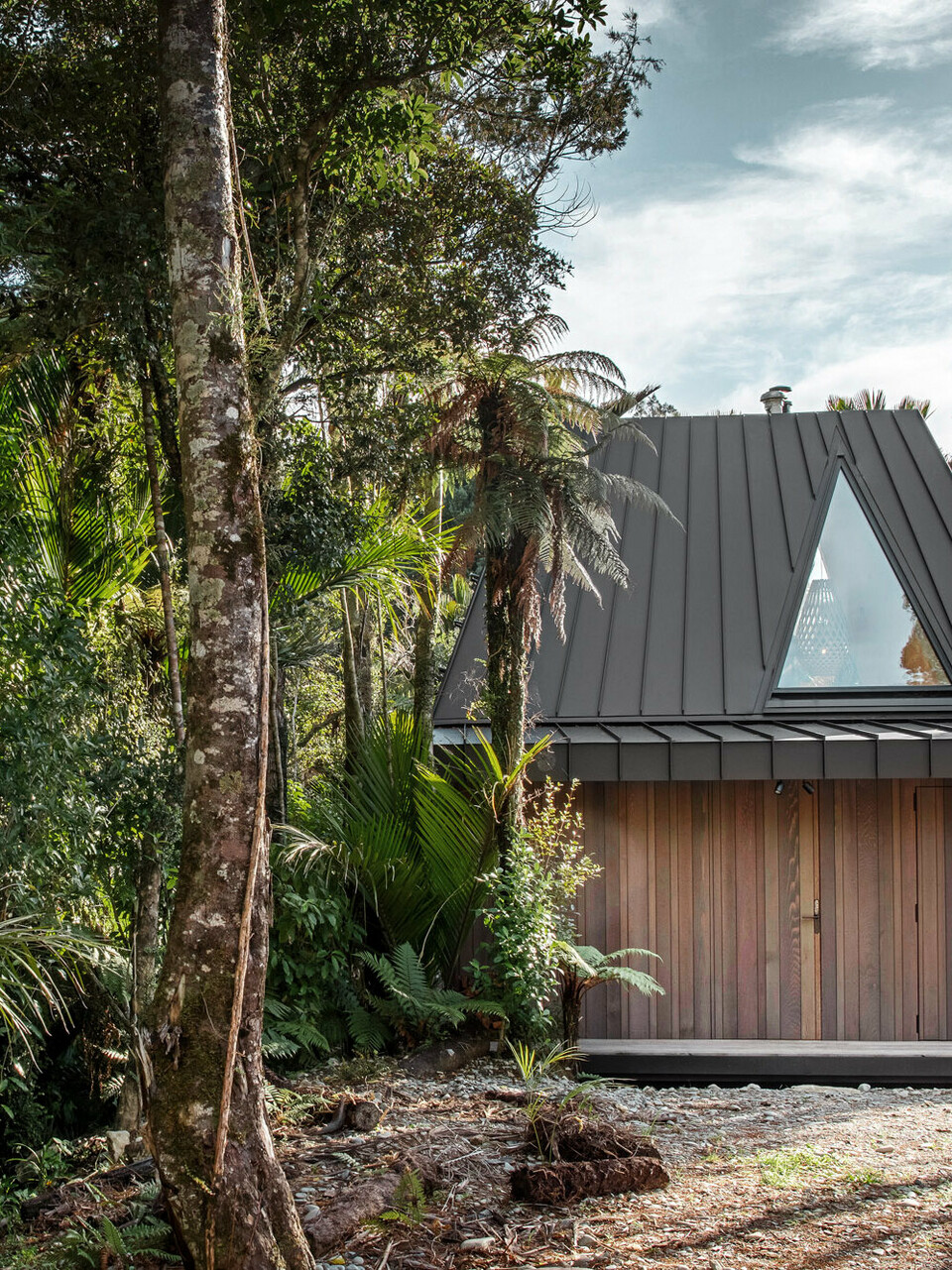 Seitlicher Blick auf das von Fabric realisierte Biwak mit dem ausgefallenen, schwarzgrauen Prefalz Dach, das sich zwischen Nikau-Palmen im Punakaiki Urwald versteckt.