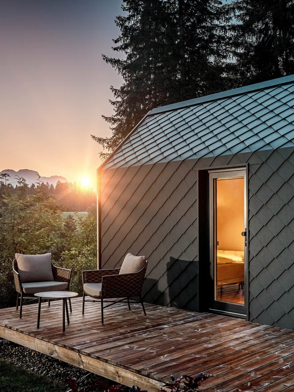 Ein minimalistisches Tiny House im Secret Koi Garden am Natterer See zeigt sich bei Sonnenuntergang mit vorgelagerter Holzterrasse und Blick in den Wald. Dach und Fassade sind mit kleinteiligen PREFA Dach‑ und Wandrauten aus Aluminium in P.10 Anthrazit ausgeführt, deren strukturierte Oberfläche das warme Licht eindrucksvoll reflektiert. Das langlebige, witterungsbeständige PREFA Aluminium schützt die Architektur zuverlässig und schafft einen markanten Kontrast zu Holz, Natur und sanfter Beleuchtung.
