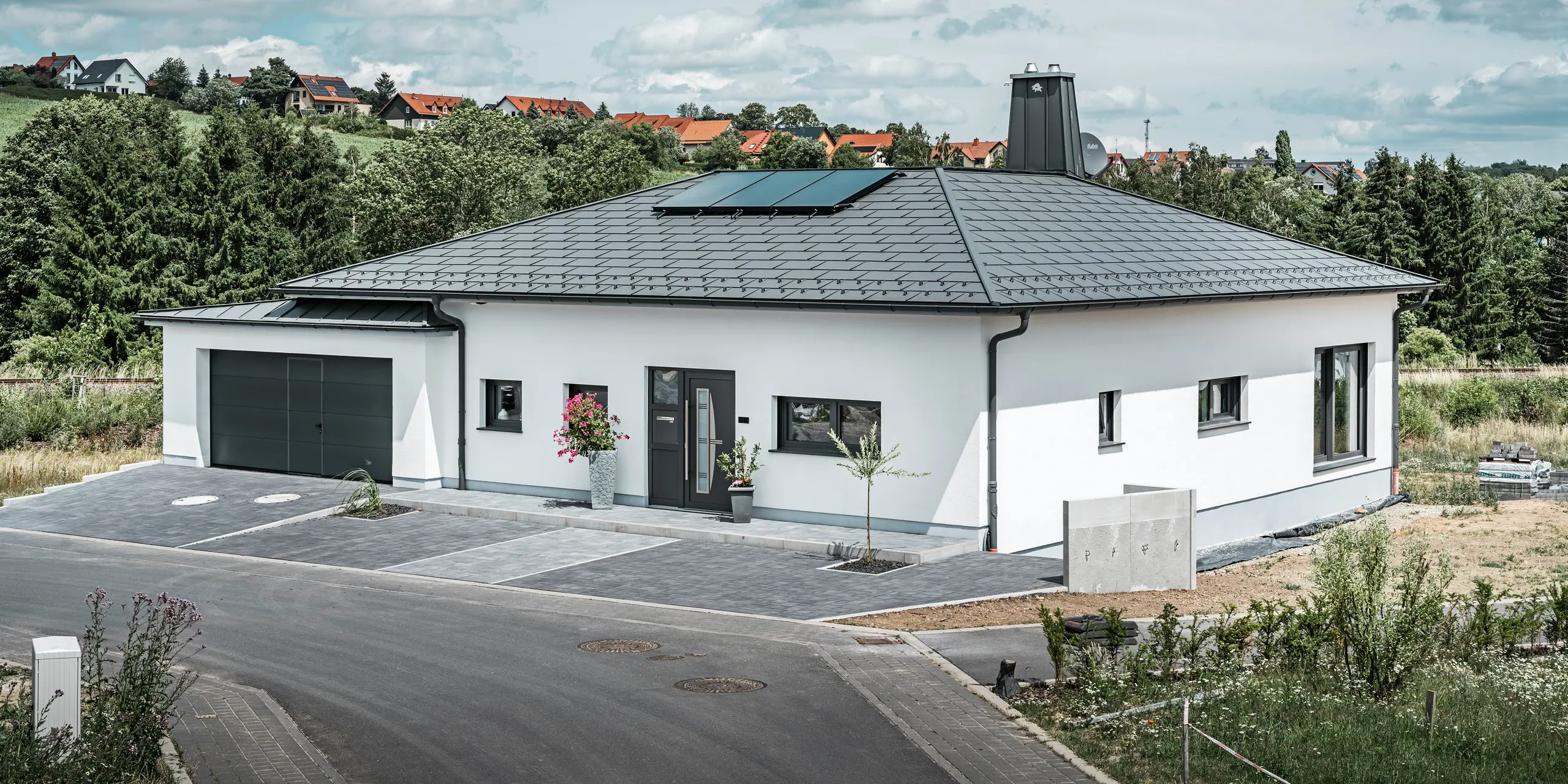Elegáns családi ház Ilmenau városában, Türingiában, kiváló minőségű PREFA tetővel, P.10 sötét szürke színben, R.16-os Classic elemmel. A garázs színében és stílusában harmonizál a PREFALZ használatával. A birtok legszembetűnőbb eleme a funkcionális kialakítású átrium és a PREFA bikával díszített kémény, amelyek hangsúlyozzák a PREFA modern dizájnját és innovatív tetőtechnológiáját. A napelemek zökkenőmentesen illeszkednek a ház fenntartható koncepciójába. A bungaló átgondolt összképét a PREFA tető-vízelvezető rendszer teszi teljessé, amely szintén sötét szürke színű.