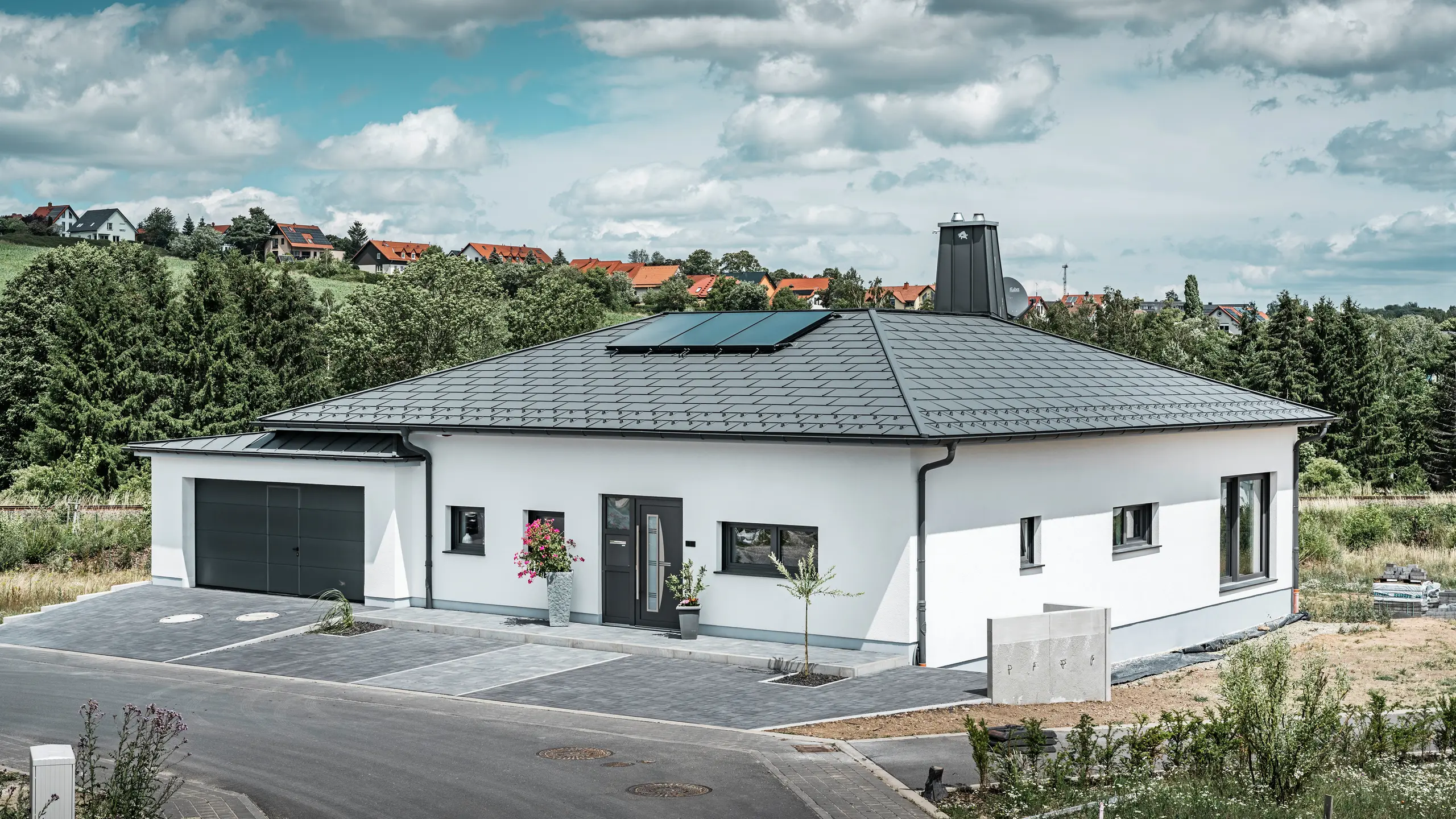 Elegáns családi ház Ilmenau városában, Türingiában, kiváló minőségű PREFA tetővel, P.10 sötét szürke színben, R.16-os Classic elemmel. A garázs színében és stílusában harmonizál a PREFALZ használatával. A birtok legszembetűnőbb eleme a funkcionális kialakítású átrium és a PREFA bikával díszített kémény, amelyek hangsúlyozzák a PREFA modern dizájnját és innovatív tetőtechnológiáját. A napelemek zökkenőmentesen illeszkednek a ház fenntartható koncepciójába. A bungaló átgondolt összképét a PREFA tető-vízelvezető rendszer teszi teljessé, amely szintén sötét szürke színű.