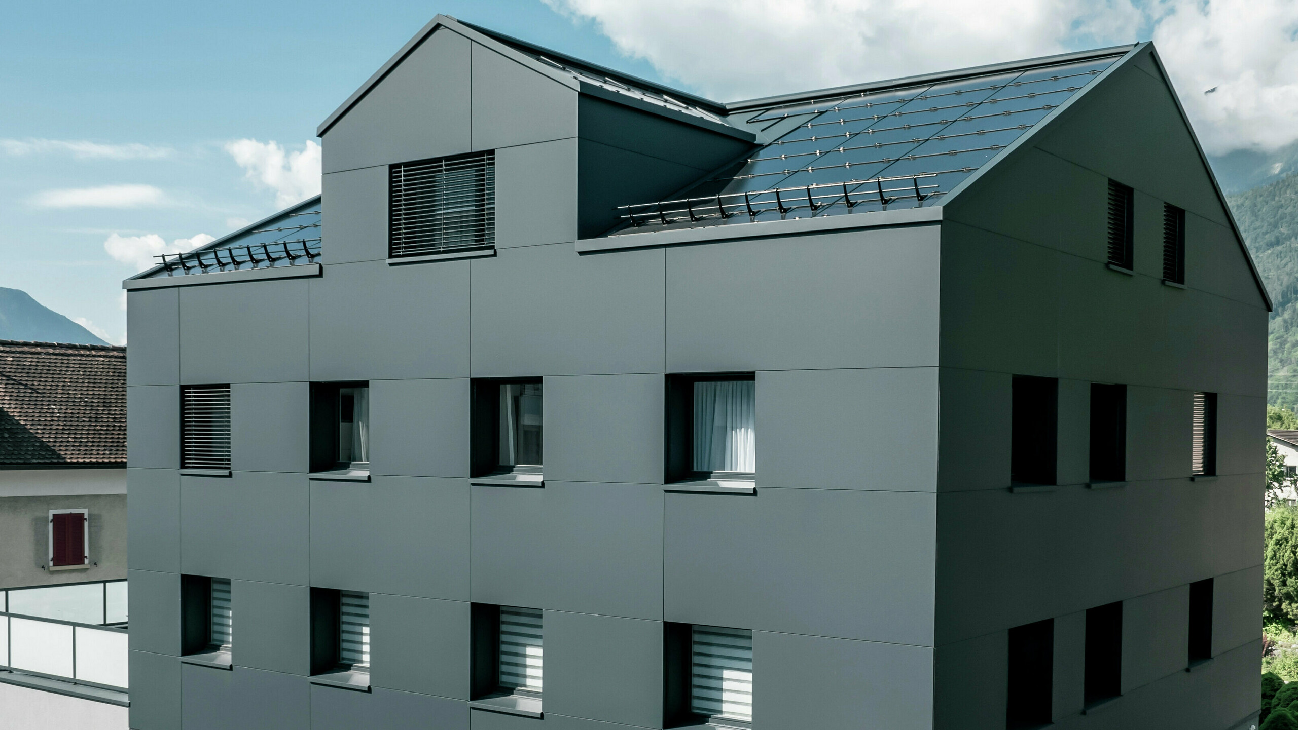 Modernisiertes Wohn- und Geschäftsgebäude nach der Fassadensanierung mit PREFABOND Aluminium Verbundplatten in Anthrazit. Die klare, elegante Linienführung der neuen Fassade verleiht dem Gebäude ein modernes Erscheinungsbild. Im Erdgeschoss befindet sich das Architekturbüro Voser. Die Umgebung mit Bäumen und Bergen rundet die harmonische Optik ab.