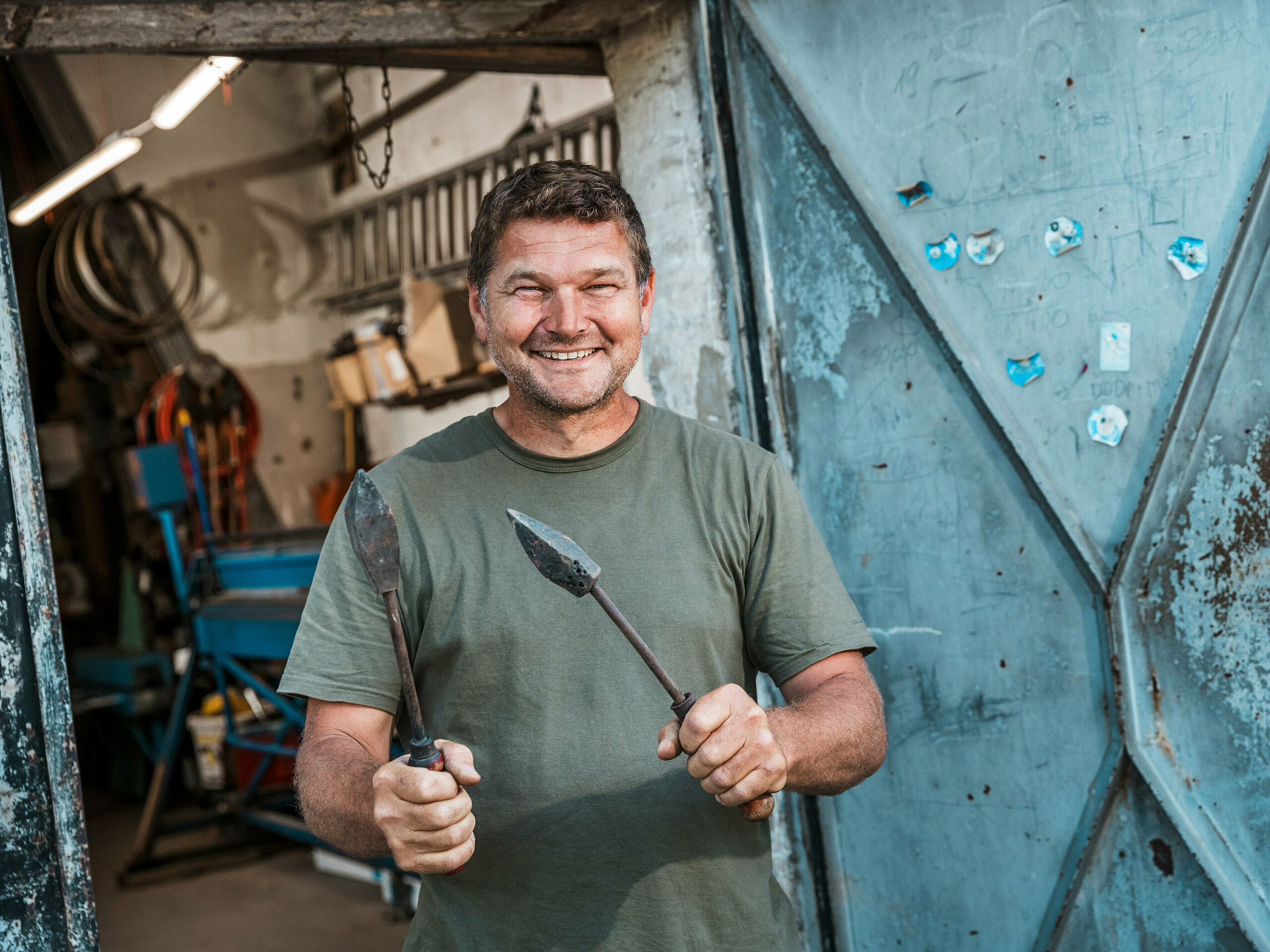 Spenglermeister Richárd Tóth in seiner Werkstatt. Der erklärte Kunsthandwerker hält traditionelles Werkzeug in den Händen und steht vor einer geöffneten Metalltür. Die Szene vermittelt authentisches Handwerk und hohe Präzision – Eigenschaften, die bei der Verarbeitung der PREFA Aluminiumprodukte für das Budapester Mehrfamilienhaus von entscheidender Bedeutung waren. Die raue Werkstattumgebung unterstreicht die handwerkliche Expertise, mit der die Fassadenelemente, die Attika und die Balkonverkleidungen aus PREFALZ in der Farbe P.10 Anthrazit gefertigt wurden. Das Bild betont die Verbindung aus Erfahrung, Materialkompetenz und langlebigen PREFA-Lösungen.