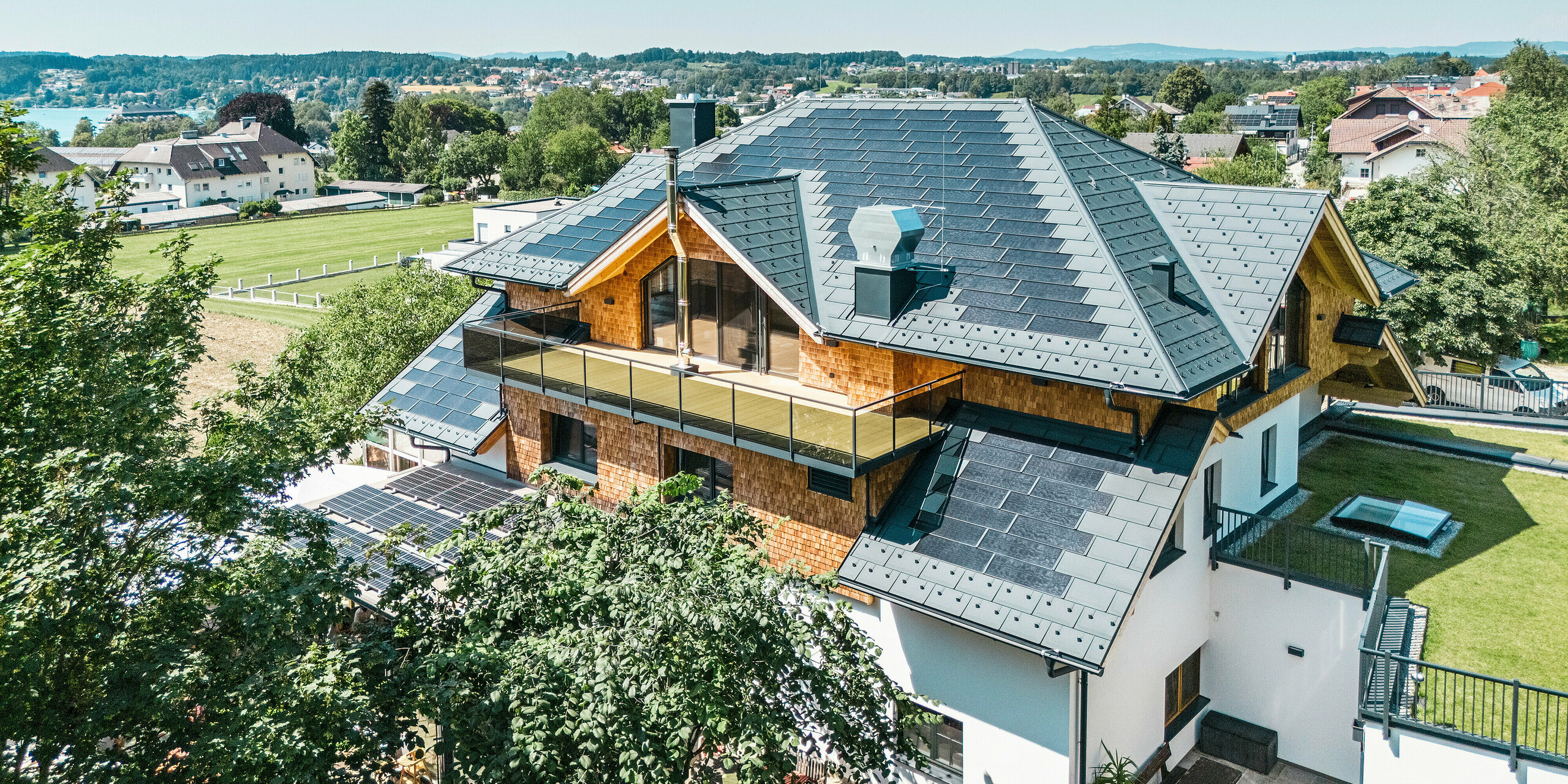 Vogelperspektive auf die Zenz'n Stub'n in Schörfling am Attersee mit neu saniertem Dach aus PREFA Solardachplatten und Dachplatten R.16 in P.10 Anthrazit. Das hochwertige Blechdach verbindet modernes Design mit nachhaltiger Energiegewinnung. Der großzügige Balkon und die harmonisch integrierte Dachentwässerung unterstreichen das stilvolle Erscheinungsbild des Gebäudes.
