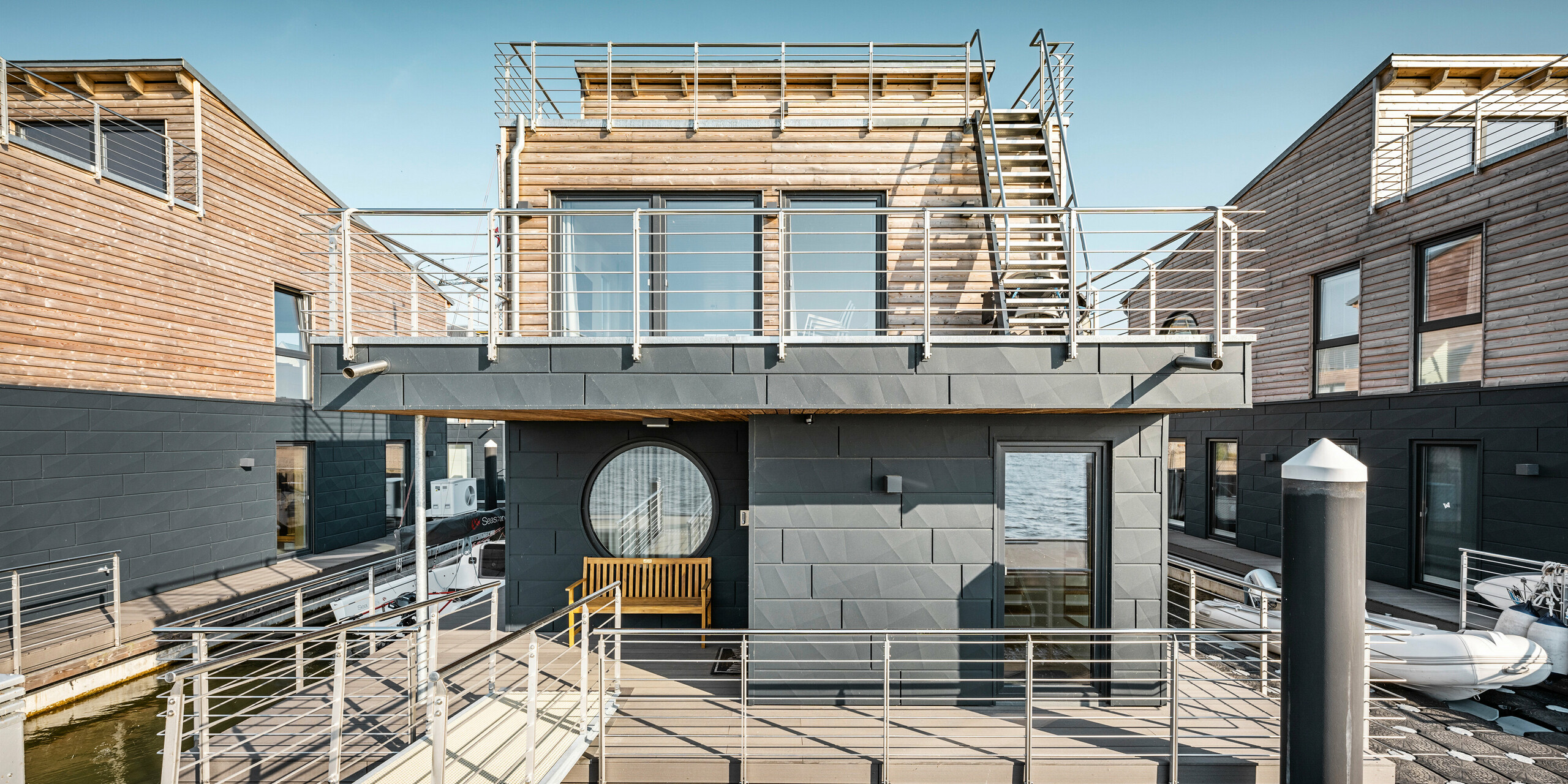 Frontansicht einer modernen Wasserhaus-Siedlung im Pionierhafen Schleswig, gekennzeichnet durch die Verwendung von nachhaltigen Materialien wie Holz und Aluminium. Die strukturierten Fassadenpaneele PREFA Siding.X in P.10 Anthrazit bieten zusammen mit den großzügigen Terrassen und Geländern aus Edelstahl eine zeitgemäße Wohnästhetik. Die großen runden und eckigen Fenster dienen als Design-Highlight und bieten einen eindrucksvollen Blick auf die Schlei, während die Wasserhäuser selbst schwimmend das Ufer säumen und so ein einzigartiges Wohnerlebnis direkt am Wasser schaffen.