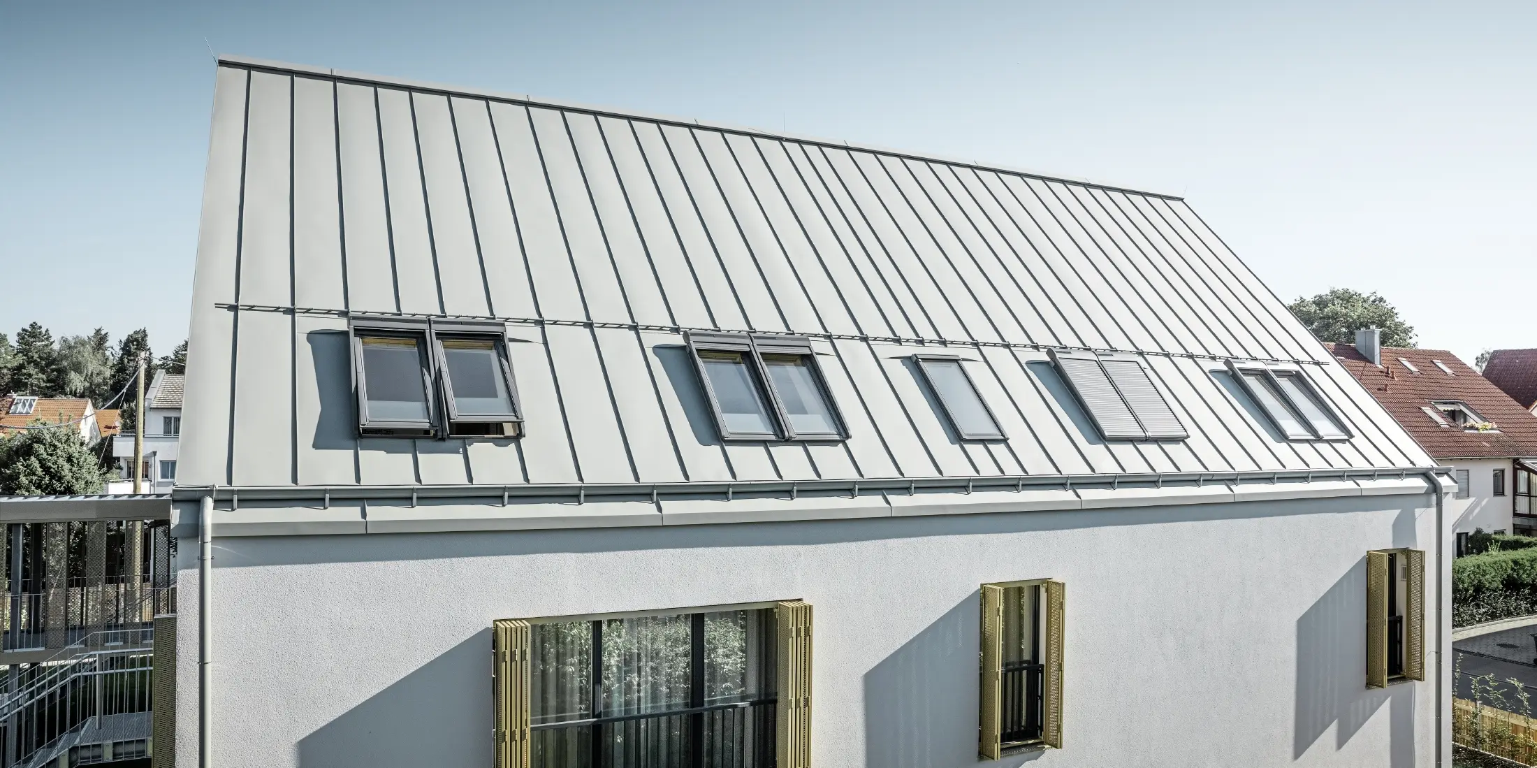 Rückansicht eines modernen Wohnheims in Augsburg mit flachem Giebel und PREFA Prefalz Zinkgrau Aluminiumdach, mehrere Dachfenster sind sichtbar, weiße Fassadenwände mit Balkonen, Umgebung mit Nachbarhäusern.