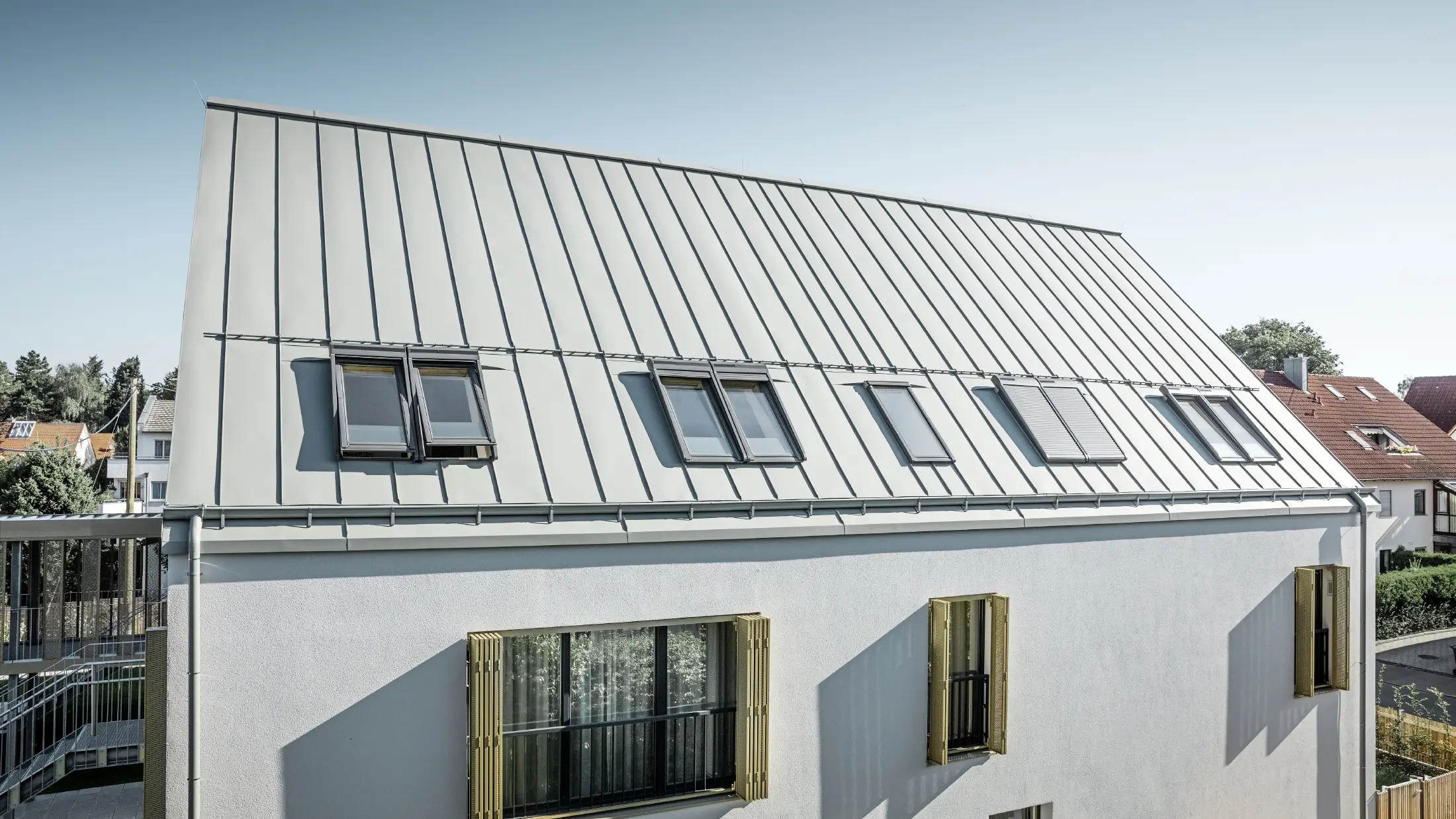 Rückansicht eines modernen Wohnheims in Augsburg mit flachem Giebel und PREFA Prefalz Zinkgrau Aluminiumdach, mehrere Dachfenster sind sichtbar, weiße Fassadenwände mit Balkonen, Umgebung mit Nachbarhäusern.