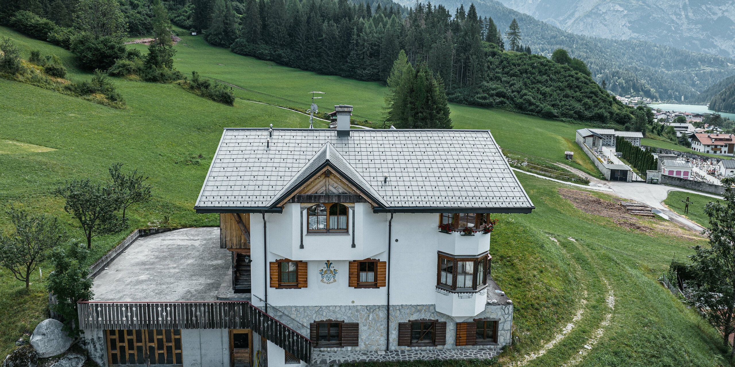 Madártávlatból az Auronzo di Cadore-i hagyományos hegyi kunyhó bejárata a PREFA P.10 világosszürke színű alumíniumcserepekkel történő tetőfelújítás után. A jellegzetes alpesi bájjal és modern bádogtetővel rendelkező épület harmonikusan illeszkedik a buja zöld tájba és a háttérben lévő lenyűgöző hegyi panorámába, a funkcionalitás és a regionális építészet sikeres kombinációját mutatva.